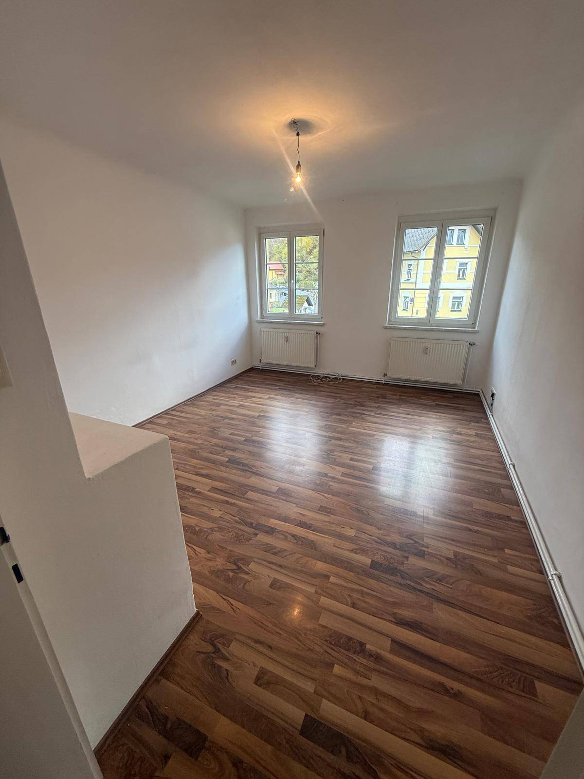 Unmöbliertes Wohnzimmer mit Holzboden und zwei Fenstern, die viel Licht hereinlassen.
