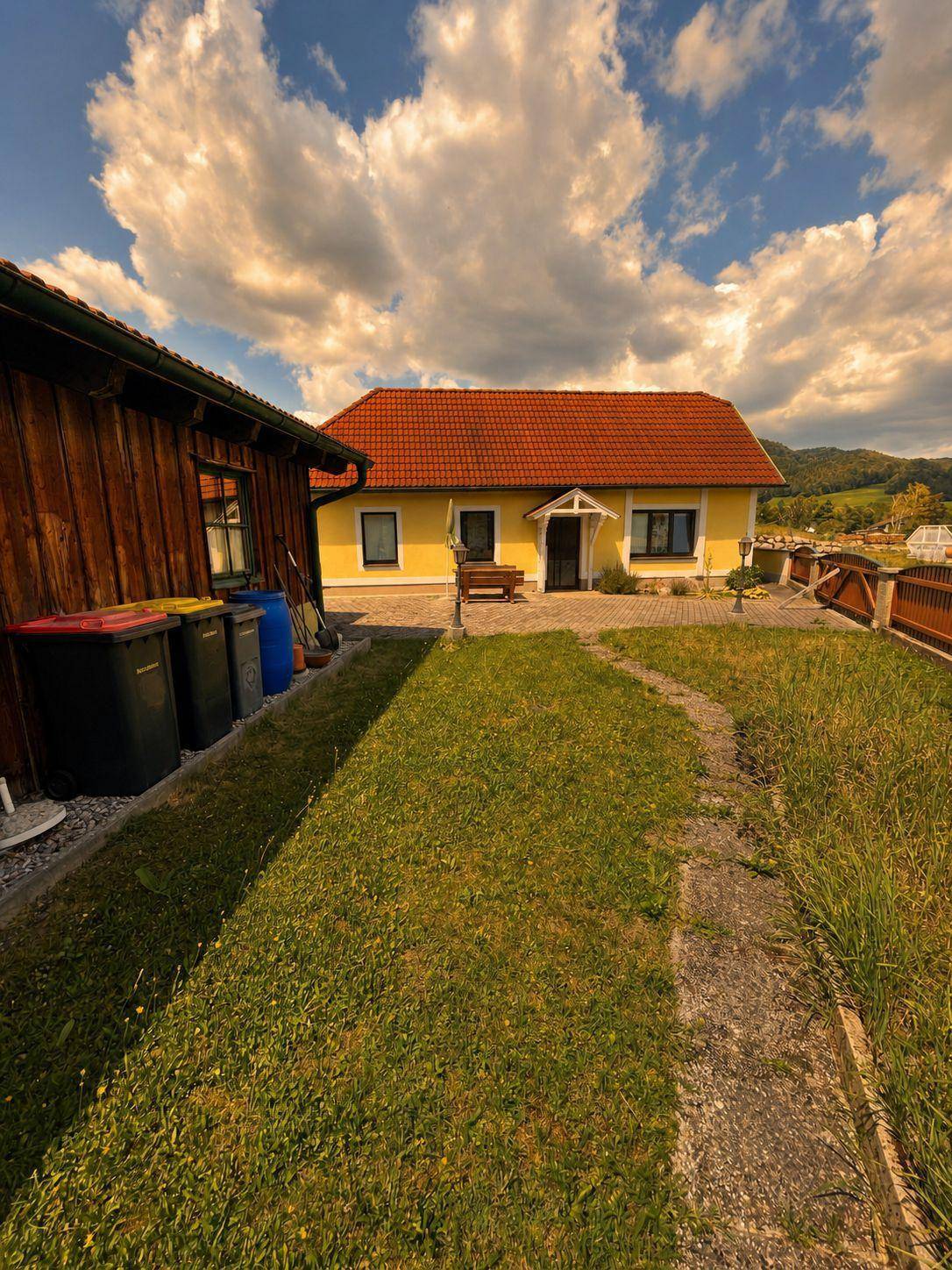 Gelbes Haus mit rotem Ziegeldach und einem Gartenweg, umgeben von grüner Wiese und Bäumen.