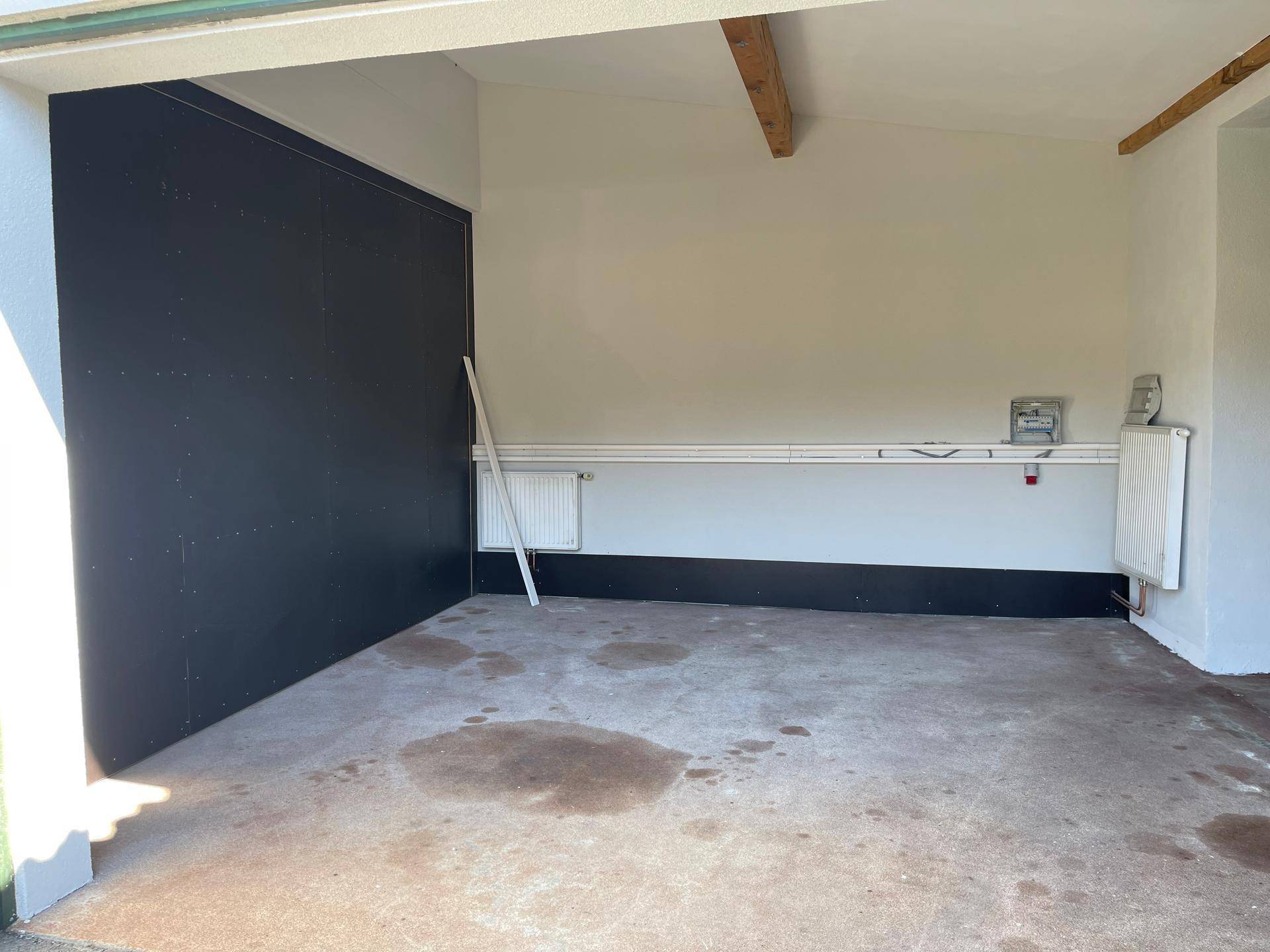 Heller Garagenraum mit Betonboden, Heizkörpern und einer dunklen Wandverkleidung, die auf Renovierungsbedarf hinweist.