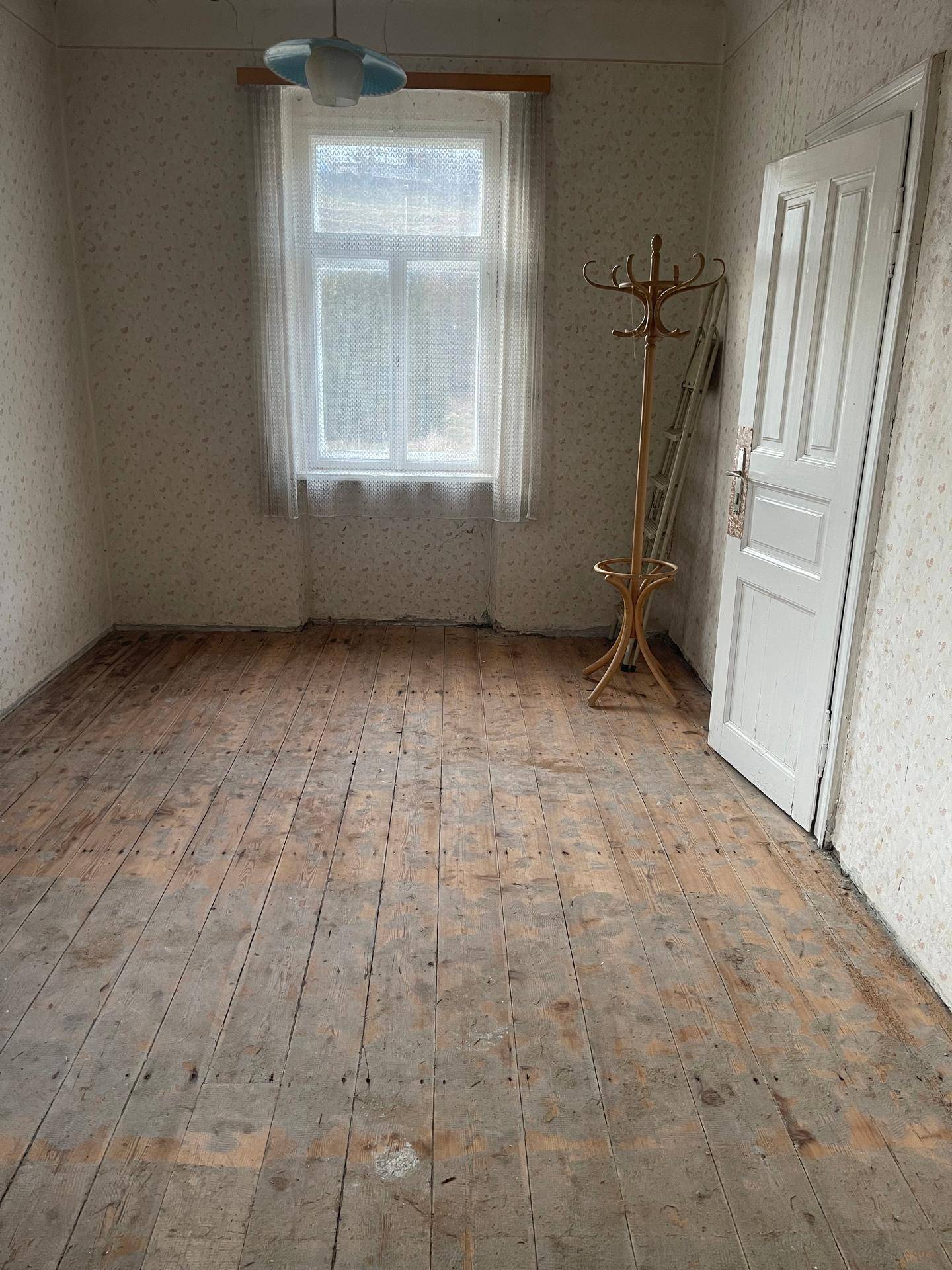 Kleines Zimmer mit Holzplankenboden, einem Fenster und alter Tapete, bereit zur Renovierung.