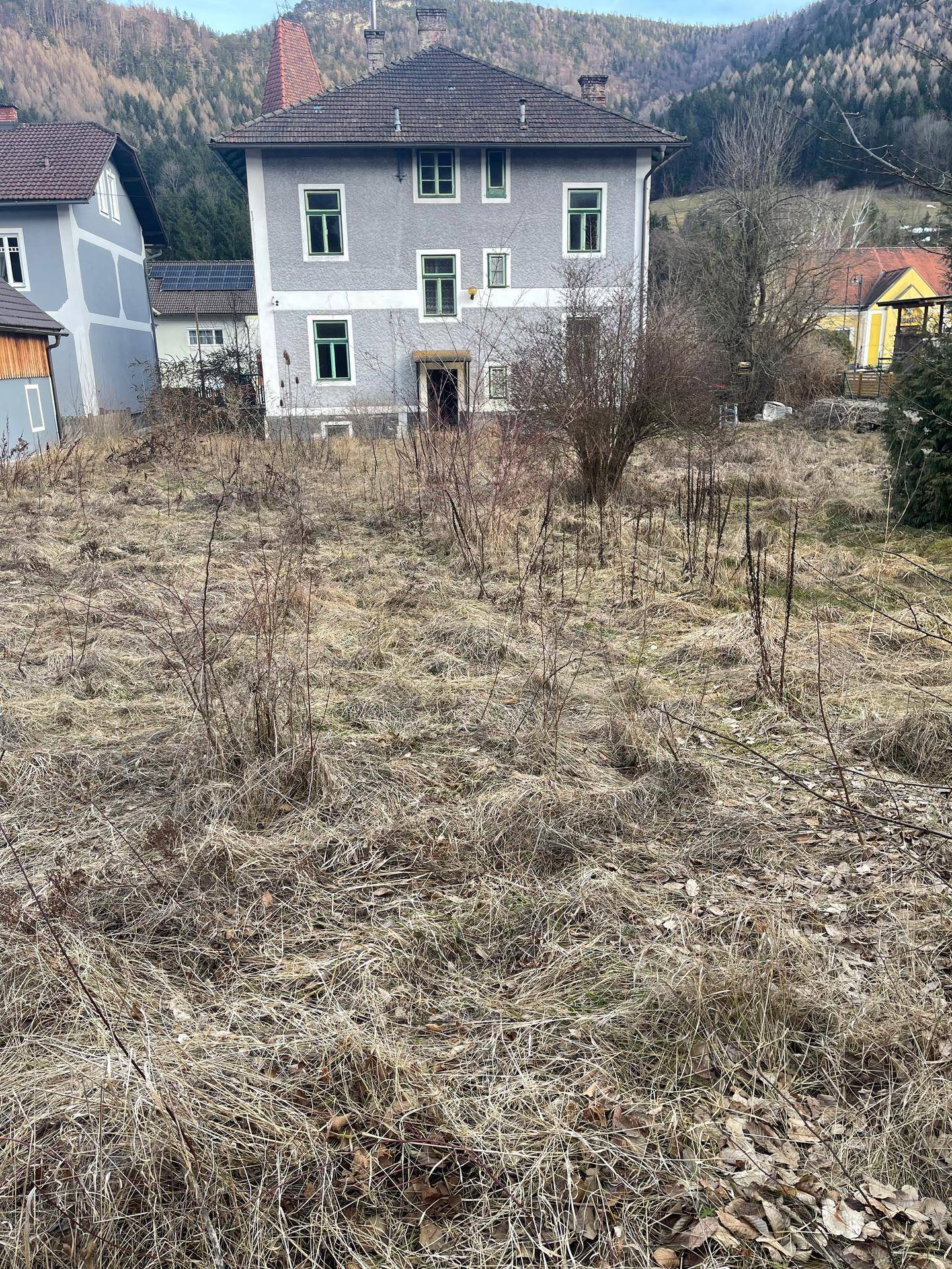 Ansicht eines grauen Gebäudes mit grünen Fenstern, umgeben von einem verwilderten Grundstück und Hügeln.