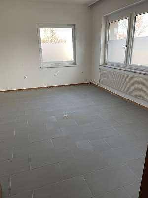 Heller, leerer Raum mit grauen Fliesen, großen Fenstern und Heizkörpern.