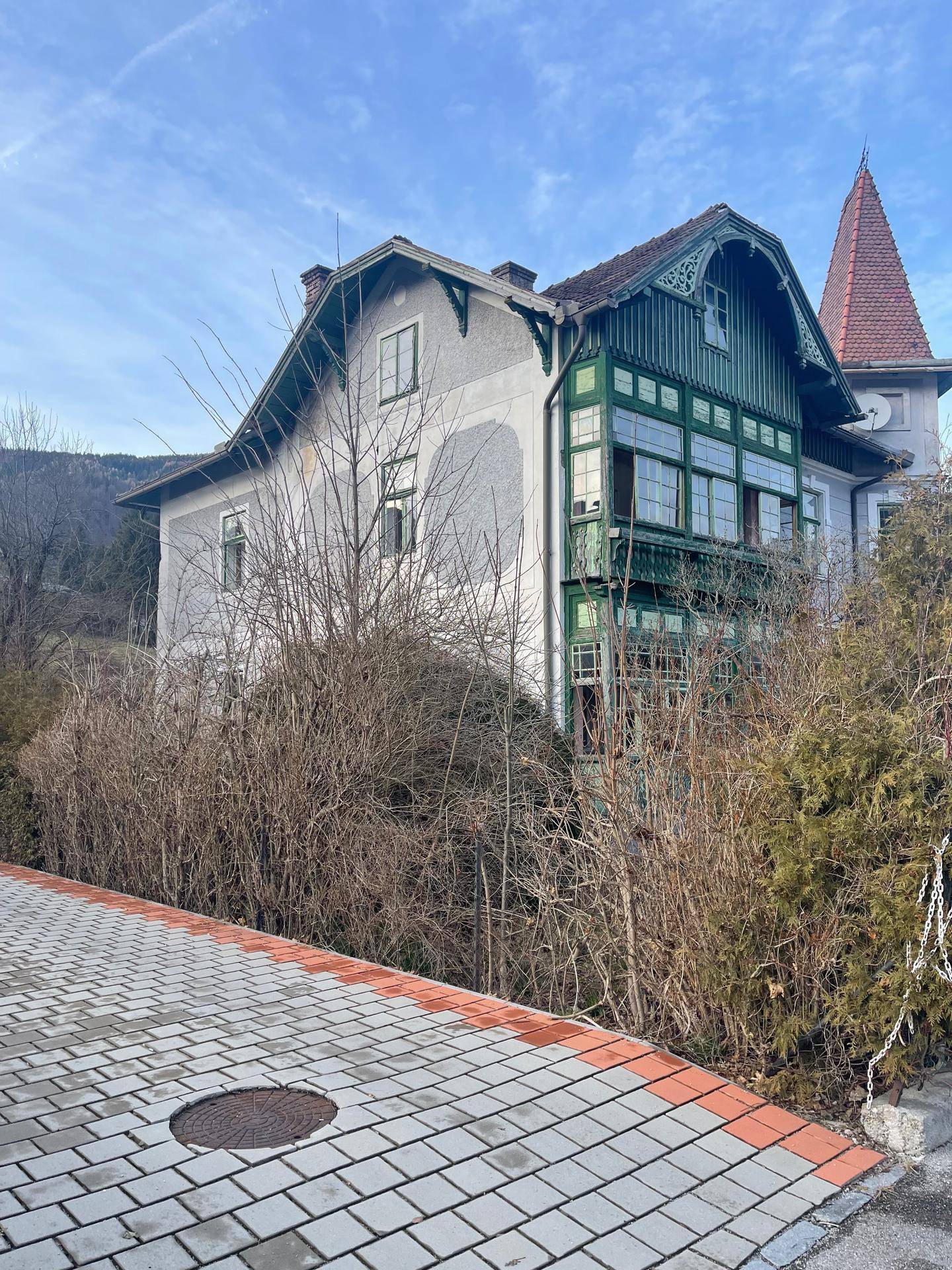 Historisches Gebäude mit grünen Holzelementen und einem Turm, umgeben von Bäumen und einem gepflasterten Weg.
