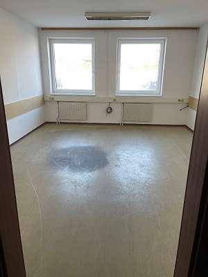 Geräumiger, leerer Raum mit hellem Boden, zwei Fenstern und Heizkörpern.