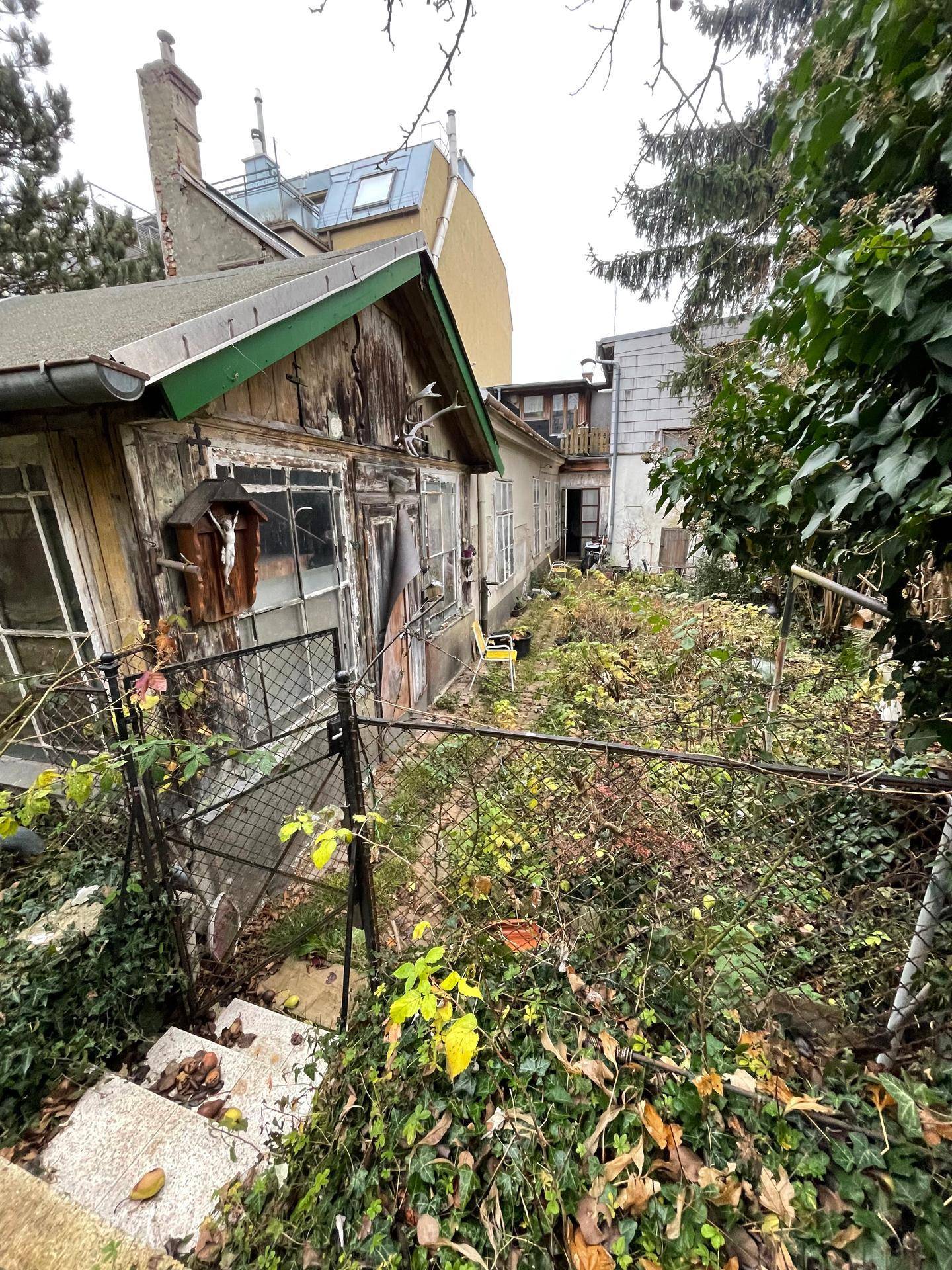 Rückseite eines baufälligen Hauses mit verwildertem Garten, altem Zaun und Treppe, umgeben von Bäumen.