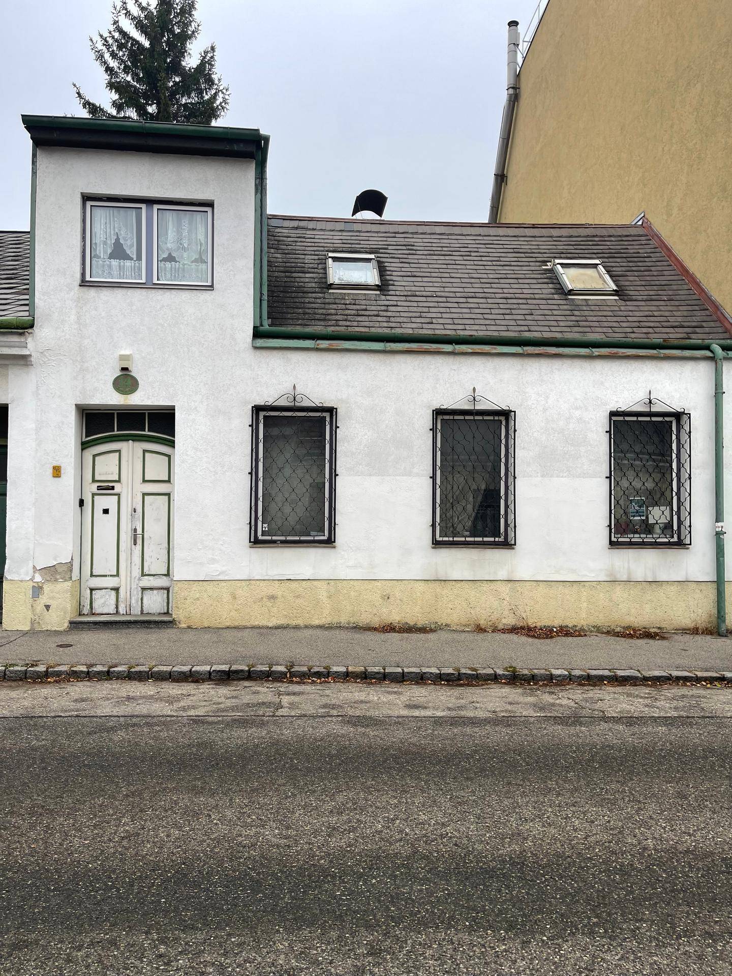 Weißes Haus mit Satteldach, kleinen Fenstern und einer Holztür, direkt an der Straße gelegen.