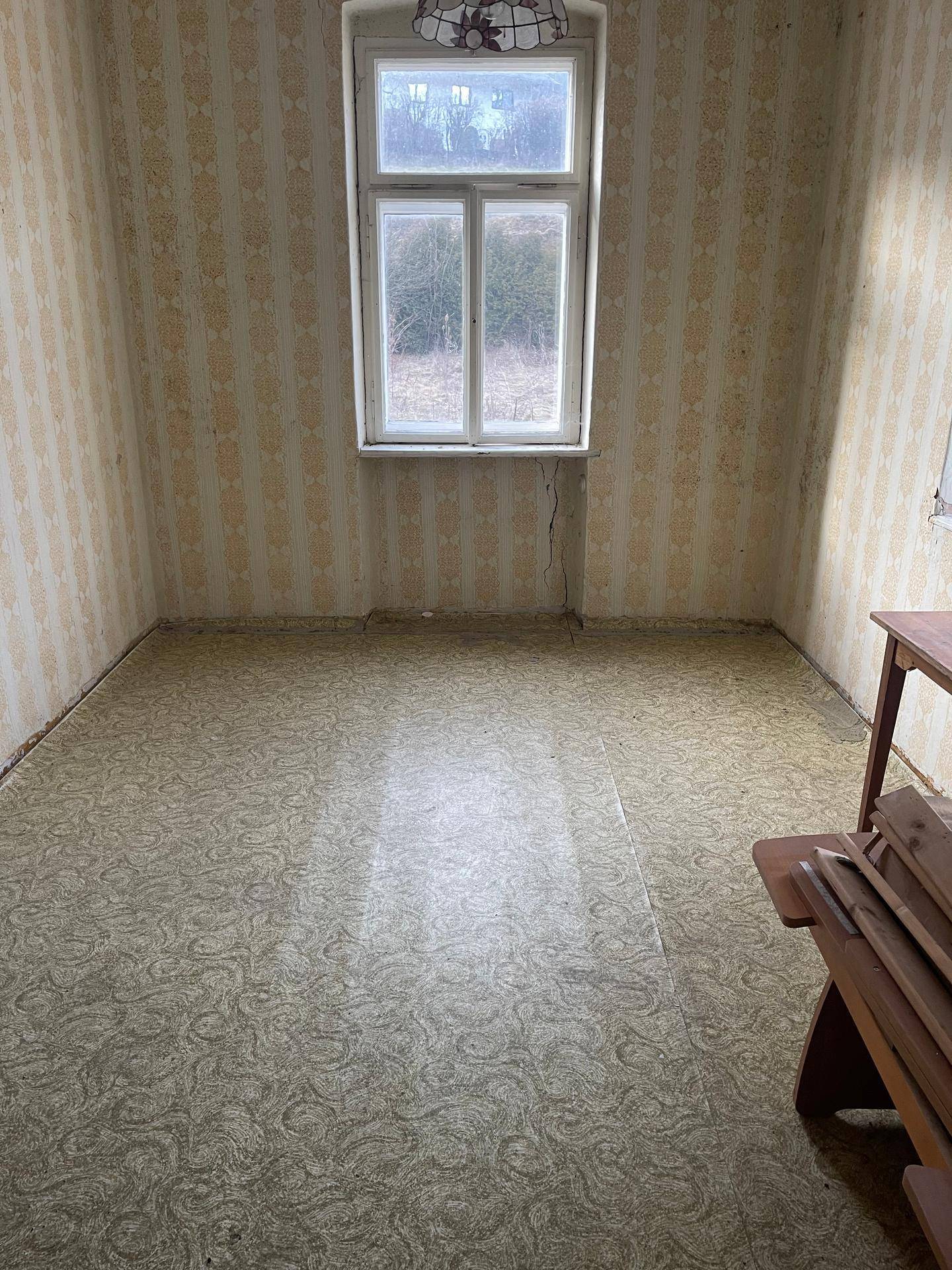 Zimmer mit gemustertem Bodenbelag, einem Fenster und alter Tapete, in einem renovierungsbedürftigen Zustand.