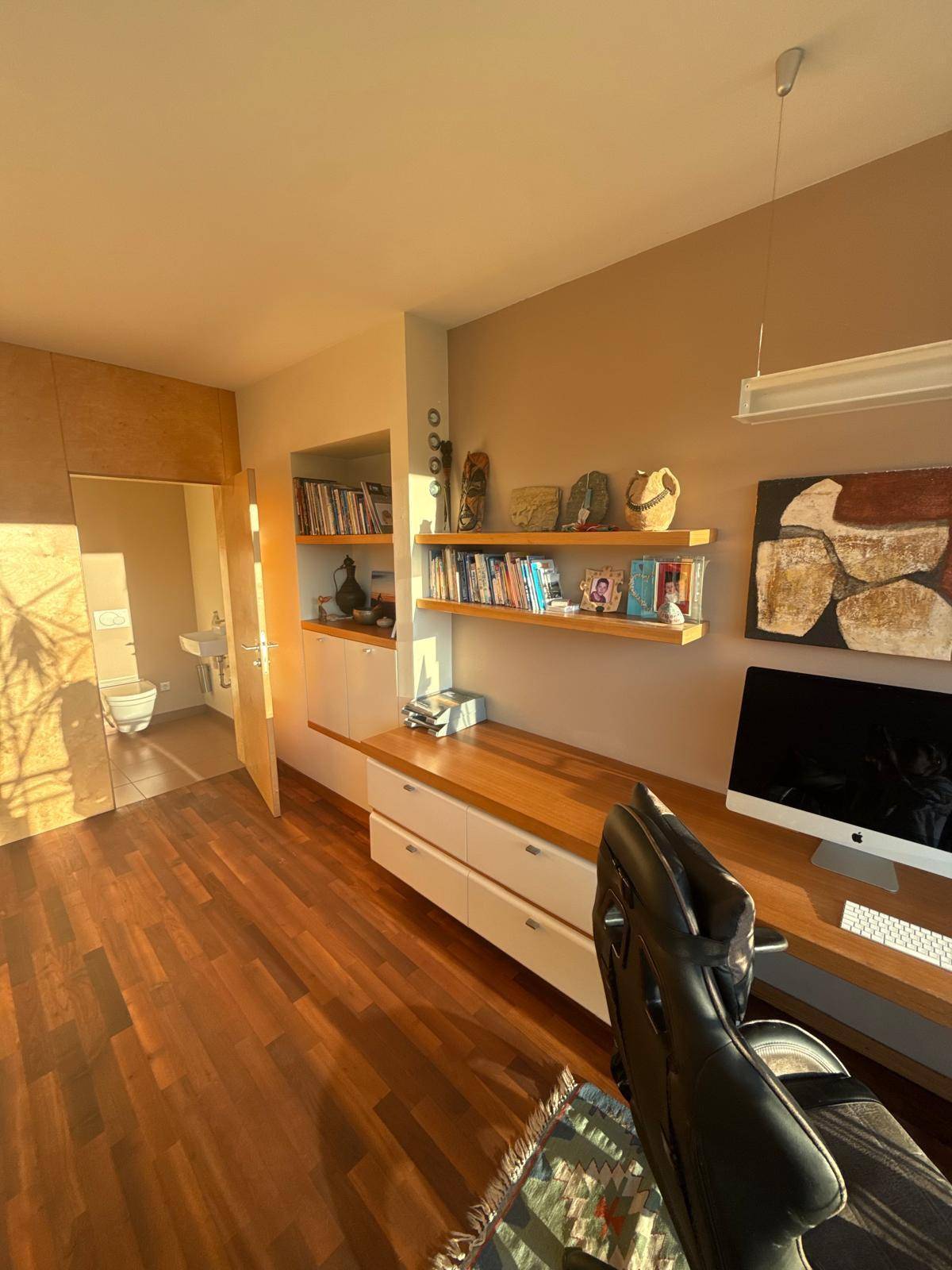 Heller Arbeitsbereich mit Holzboden, Schreibtisch und integrierten Regalen, mit Blick auf ein WC.