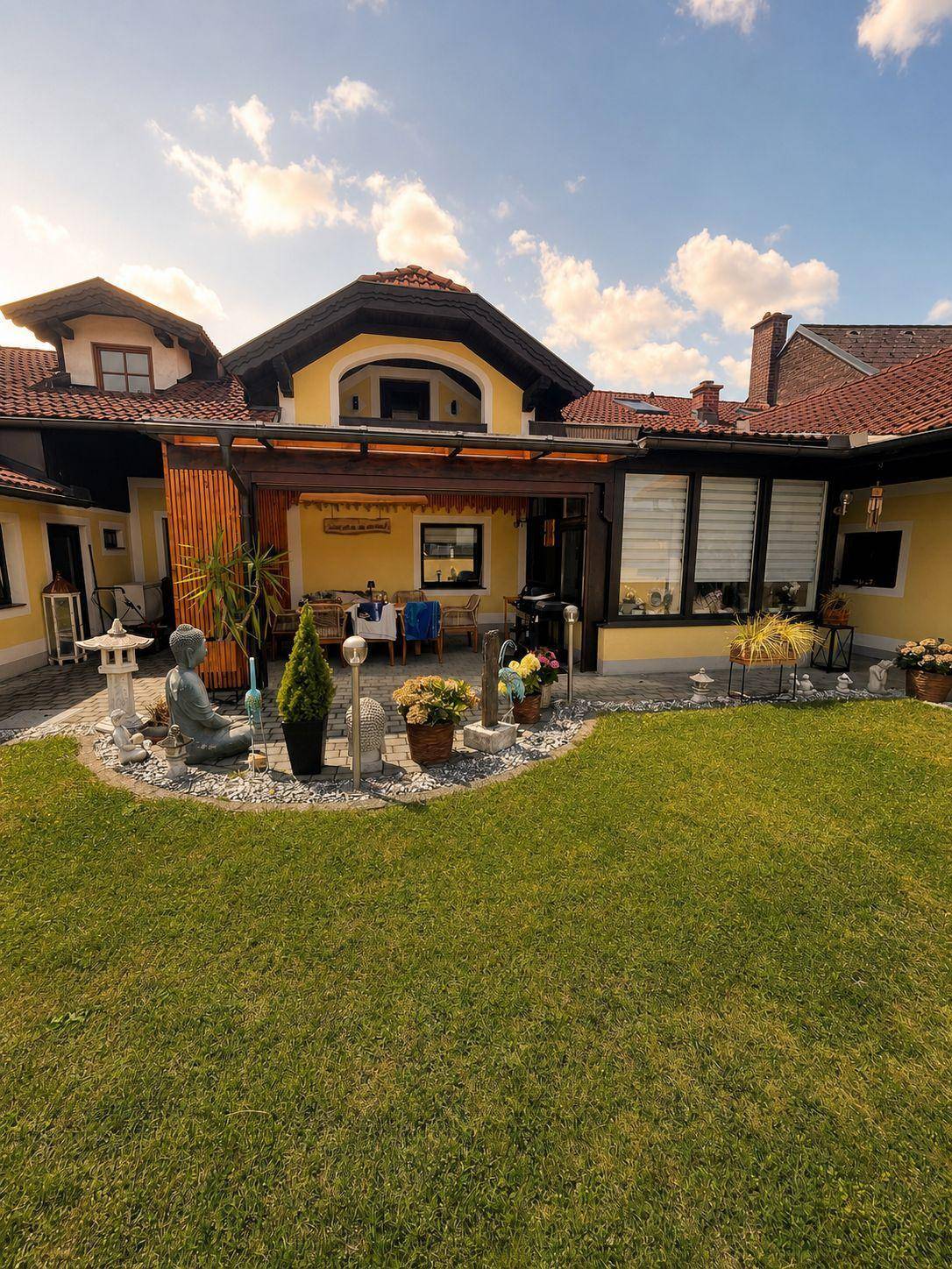 Gepflegter Garten mit Rasenfläche, Terrasse und einem gelben Haus im Hintergrund unter blauem Himmel.