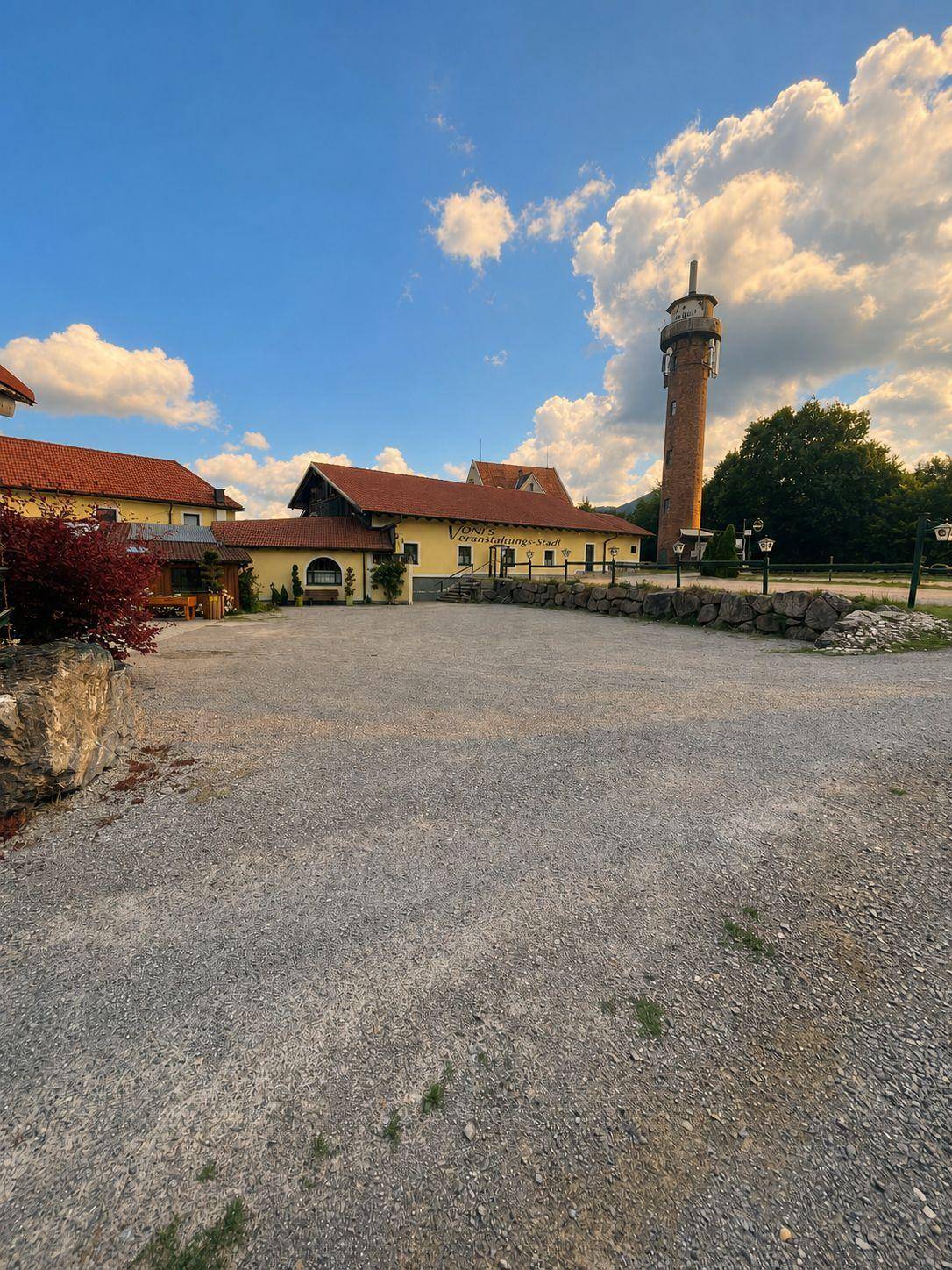 Weitläufiger Schotterparkplatz vor dem Gebäudekomplex mit Turm und grüner Umgebung.