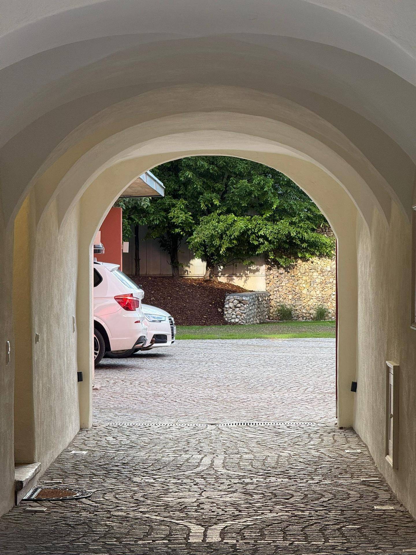 Blick durch einen Torbogen auf einen gepflasterten Innenhof mit parkenden Autos und Grünflächen.