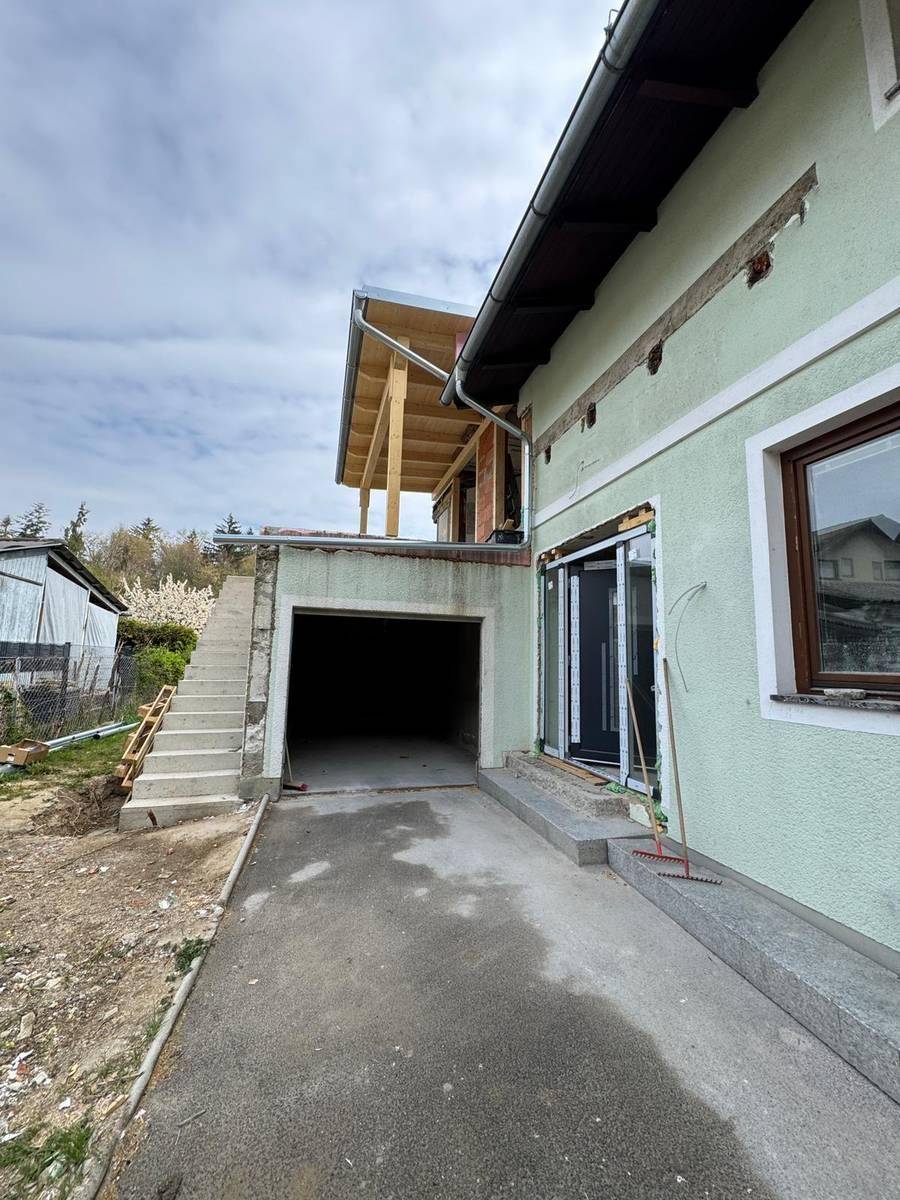 Seitenansicht des Hauses mit integrierter Garage und Außentreppe, umgeben von Baustellenmaterial.
