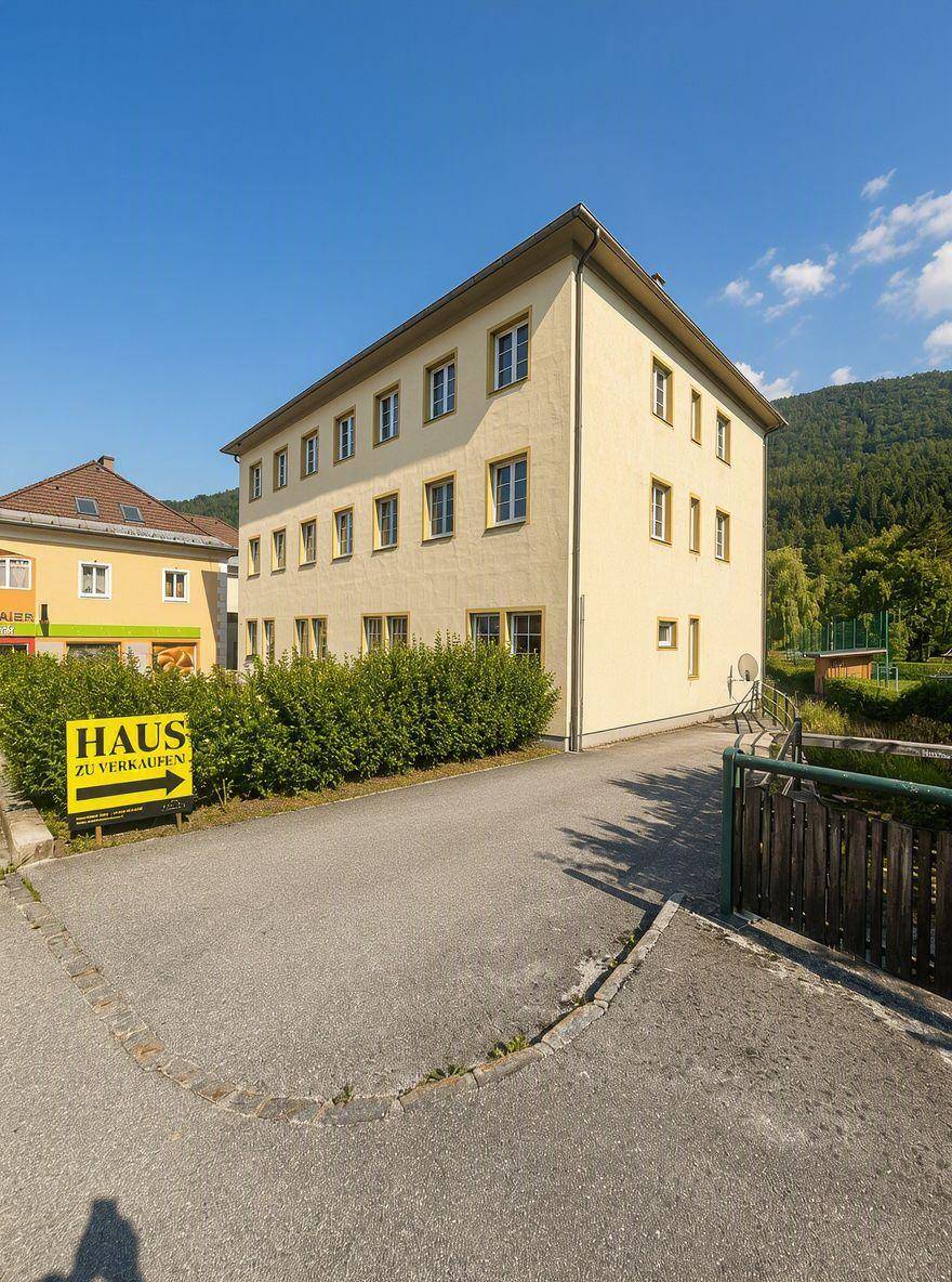 Gelbes Gebäude mit "Haus zu verkaufen" Schild und Blick auf die grüne Umgebung.