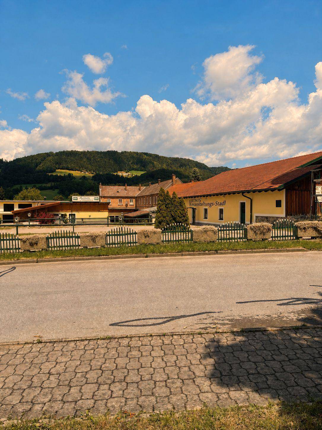 Gebäude mit gelber Fassade und rotem Dach, umgeben von grünen Hügeln und Bäumen.