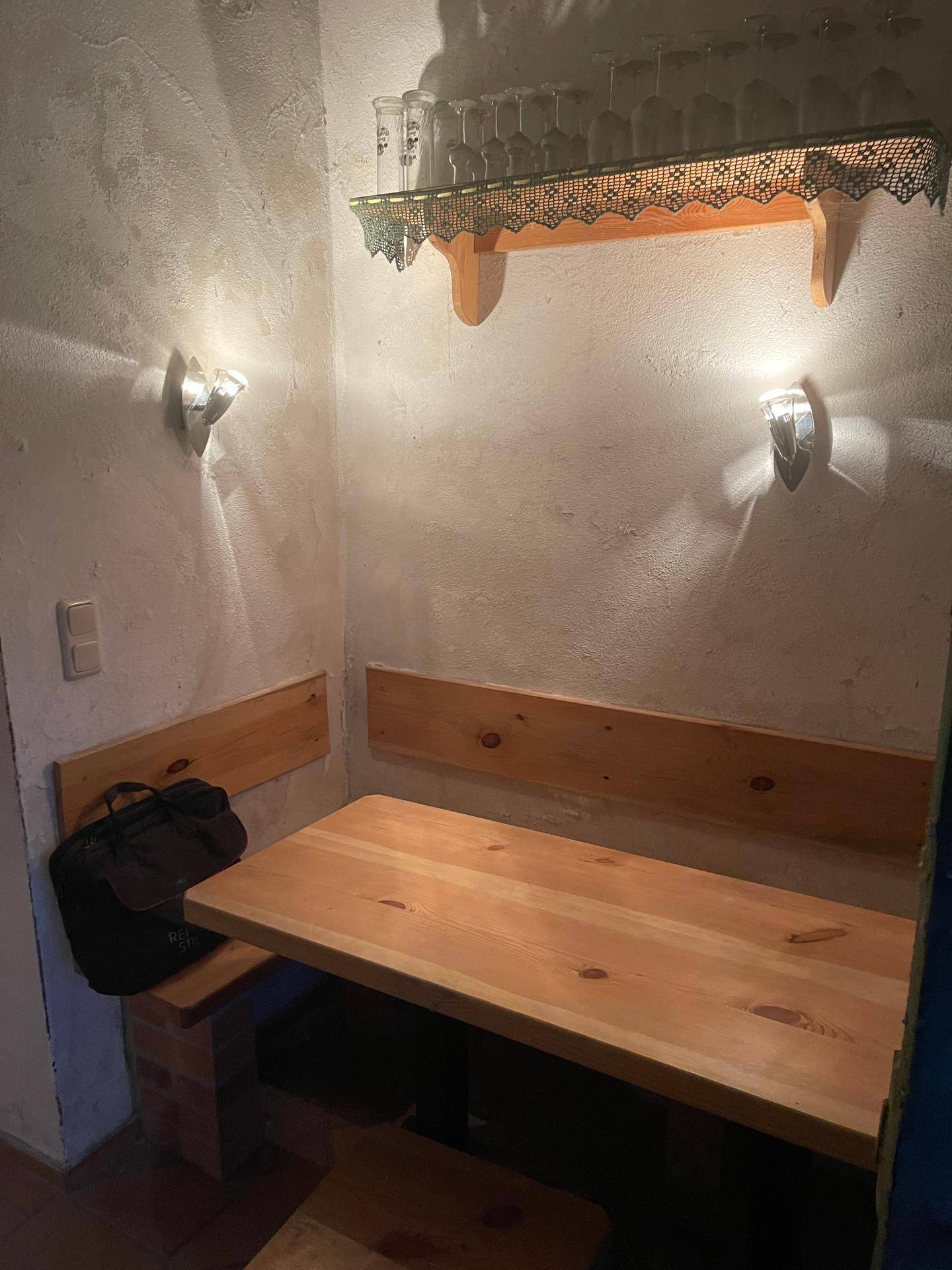 Einfacher, rustikaler Sitzbereich mit Holztisch und Bänken in einem Kellerraum, beleuchtet von Wandleuchten.