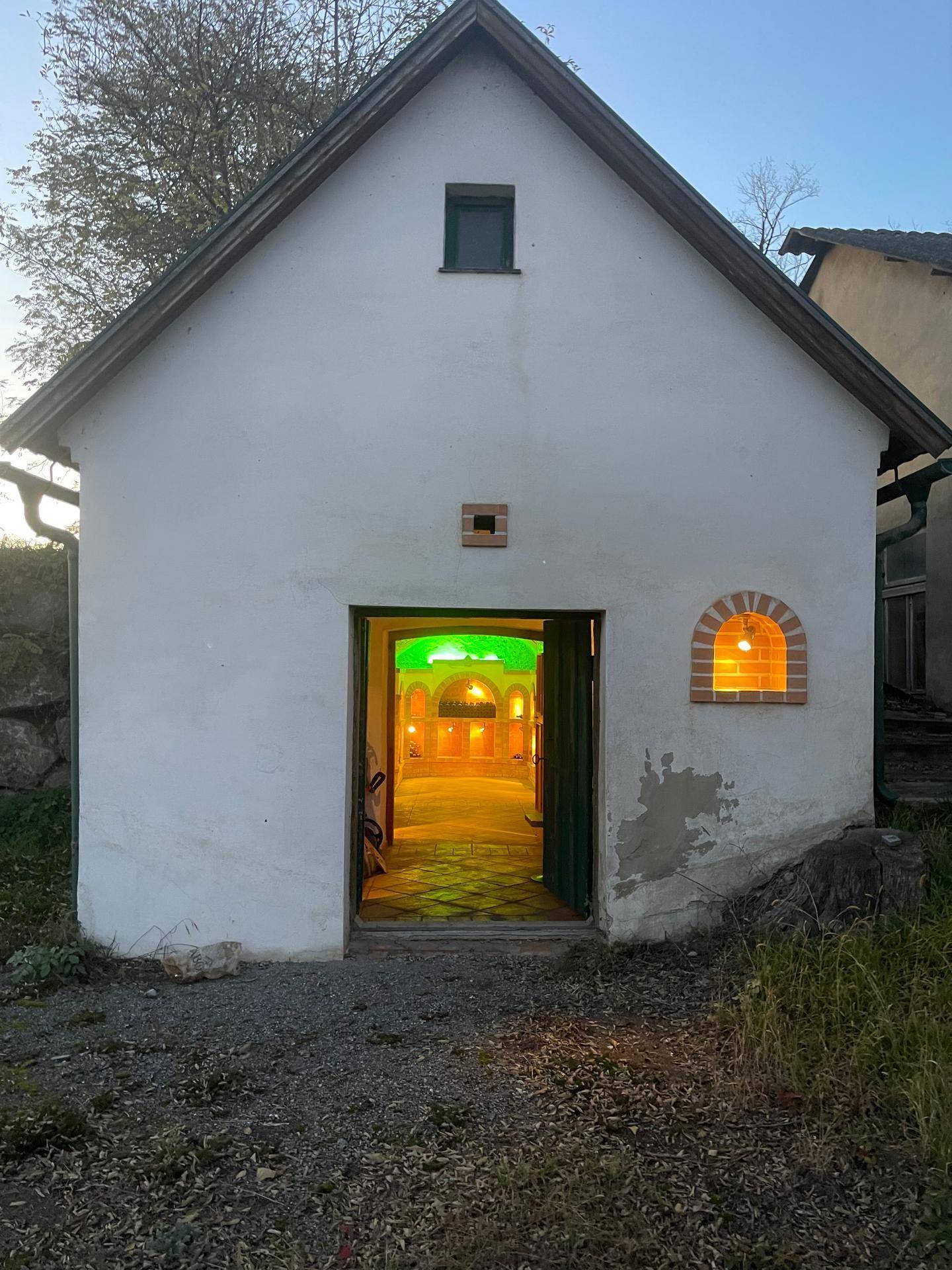 Traditionelles Kellergebäude mit weißer Fassade und grün beleuchtetem Eingang, umgeben von natürlicher Landschaft.