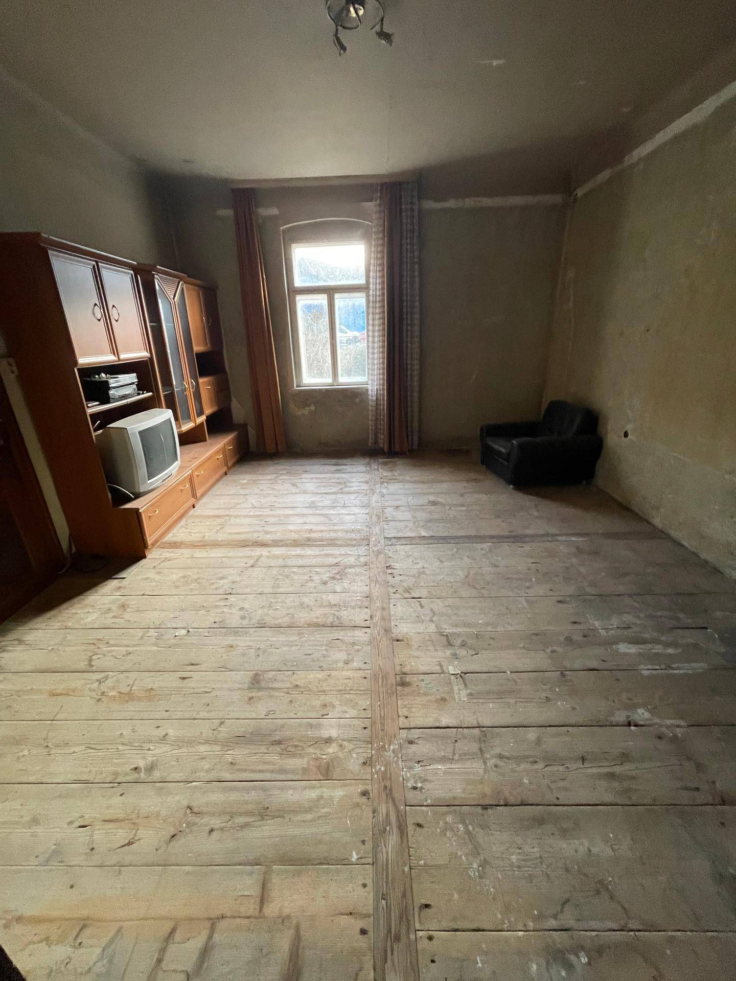Wohnzimmer mit breiten Holzplanken, einem Fenster und einem alten Schrank, der Renovierung bedarf.