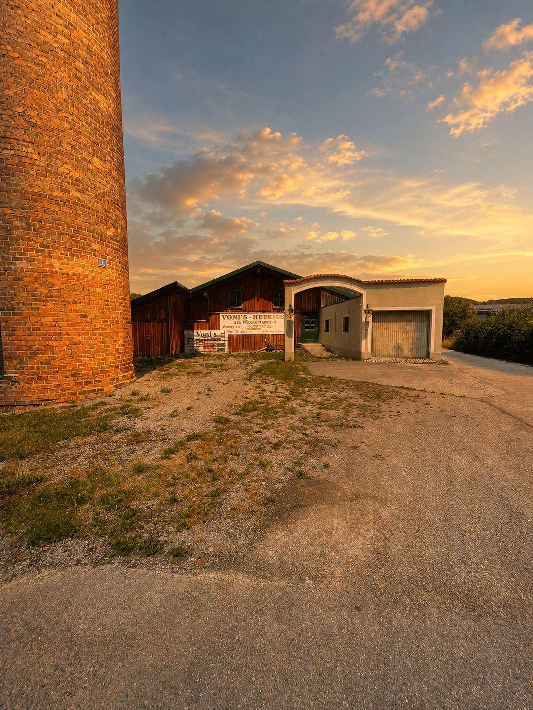 Ansicht eines Holzgebäudes mit Torbogen und Garagentor, neben dem hohen Turm bei Sonnenuntergang.