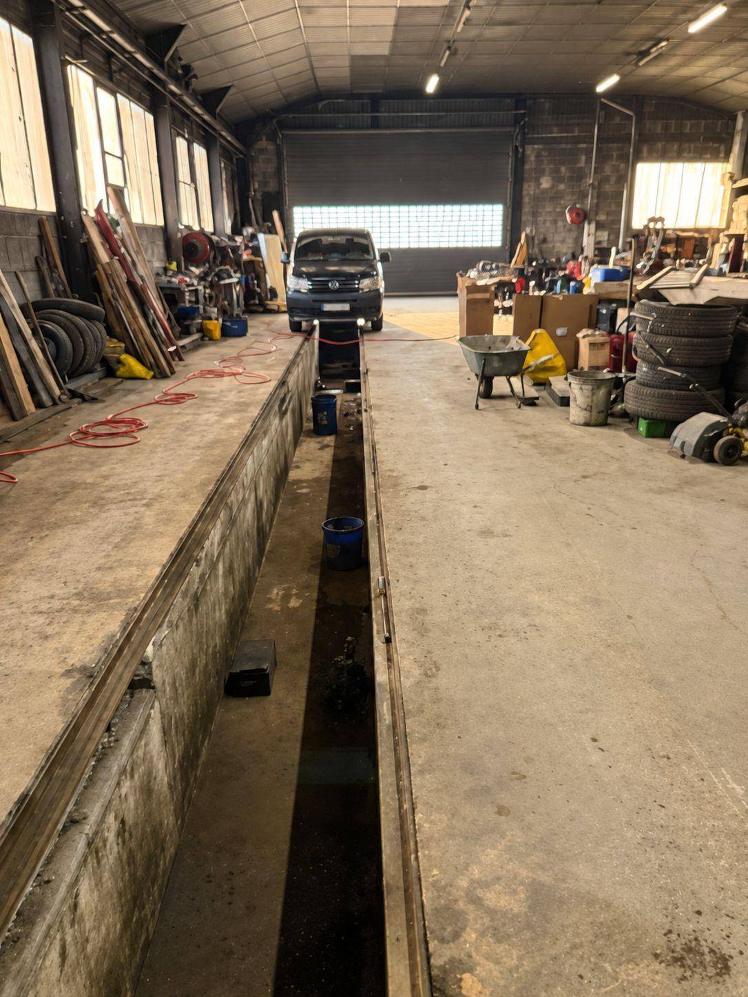 Innenansicht einer Werkstatt mit einer Wartungsgrube, einem geparkten Transporter und verschiedenen Werkzeugen.