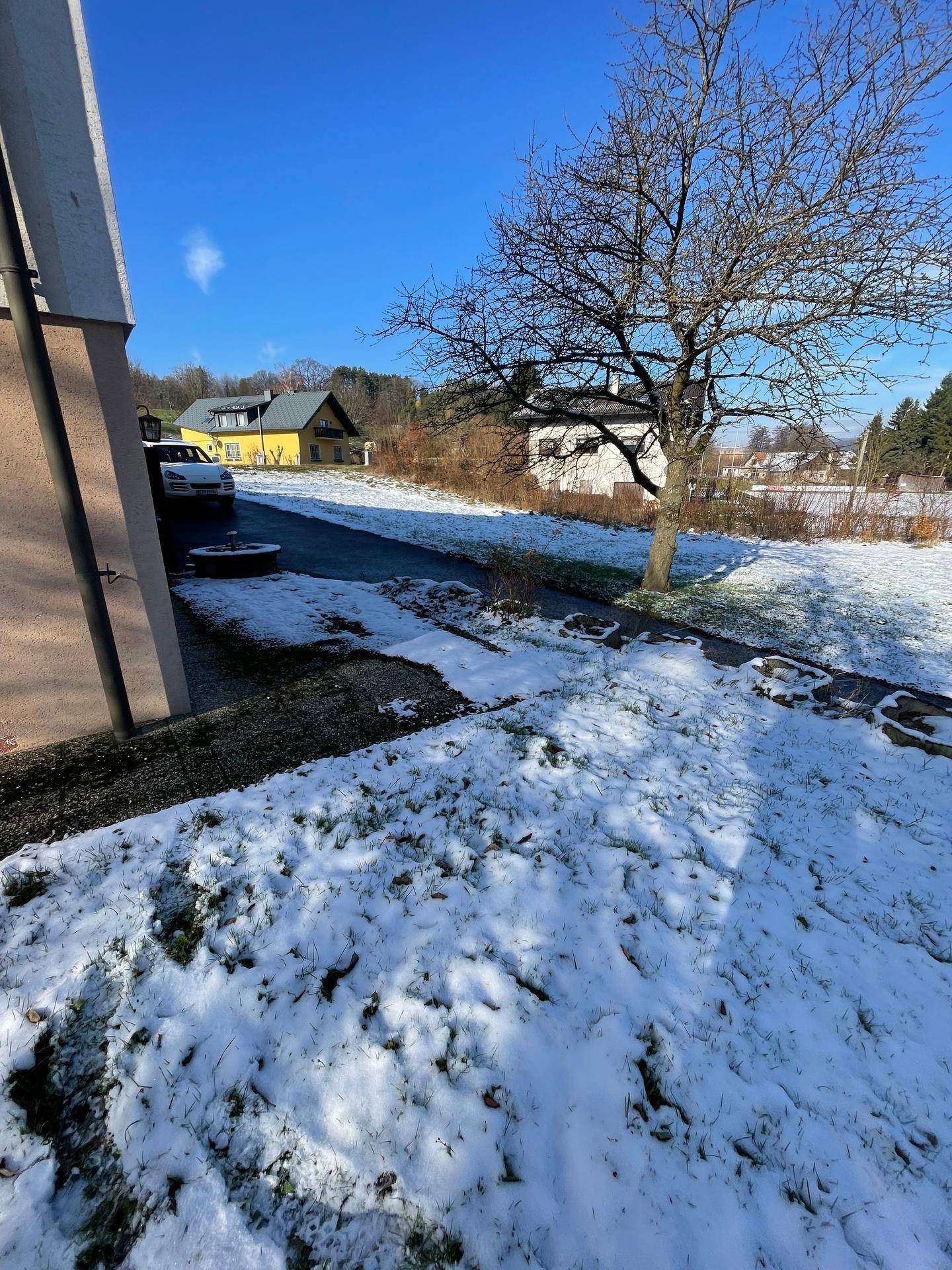 Schneebedeckter Gartenweg neben einem Haus mit kahlen Bäumen und weiteren Gebäuden im Hintergrund.