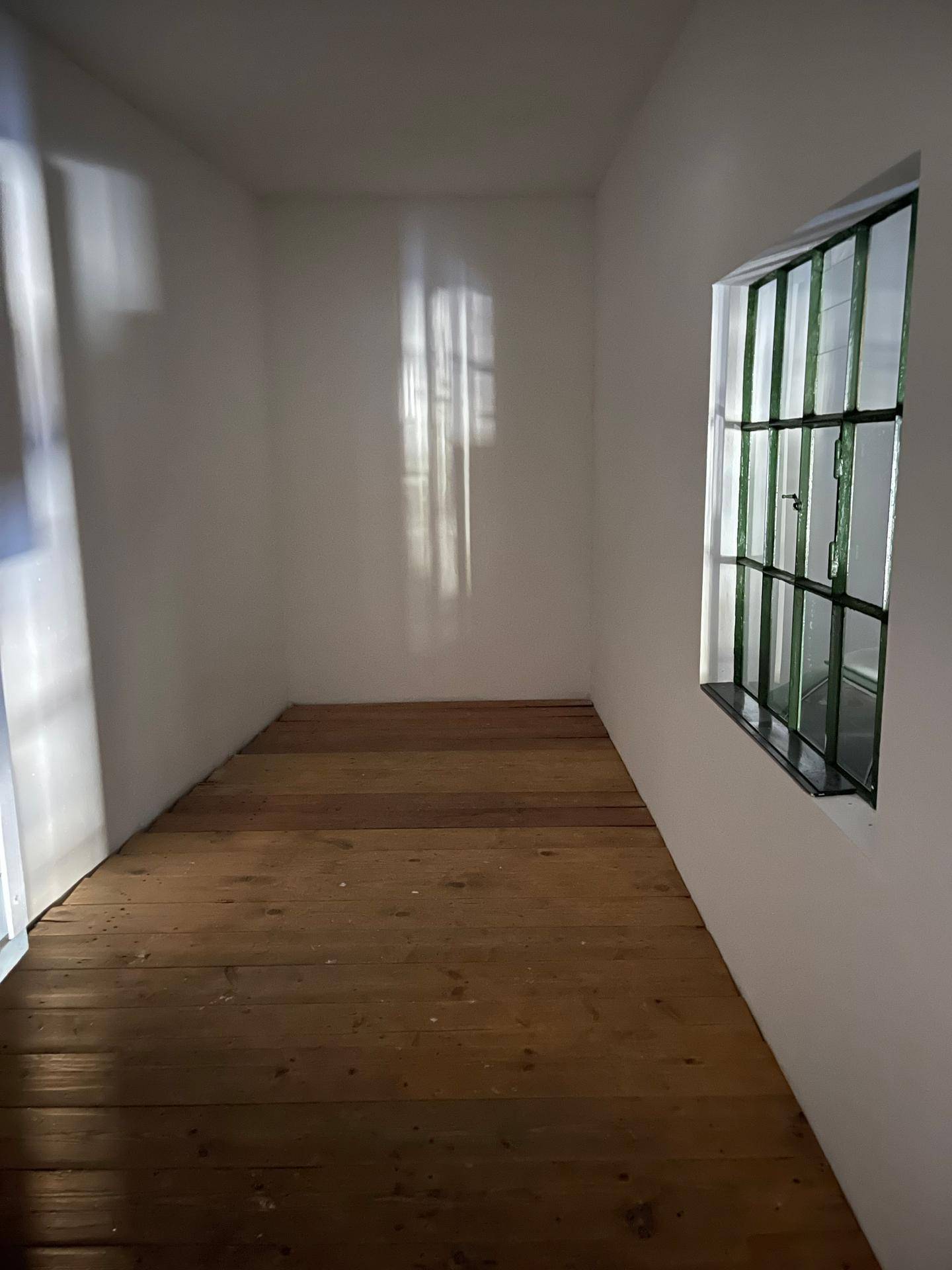 Kleiner, leerer Raum mit Holzdielenboden und einem Fenster mit grünen Gitterstäben, das natürliches Licht hereinlässt.