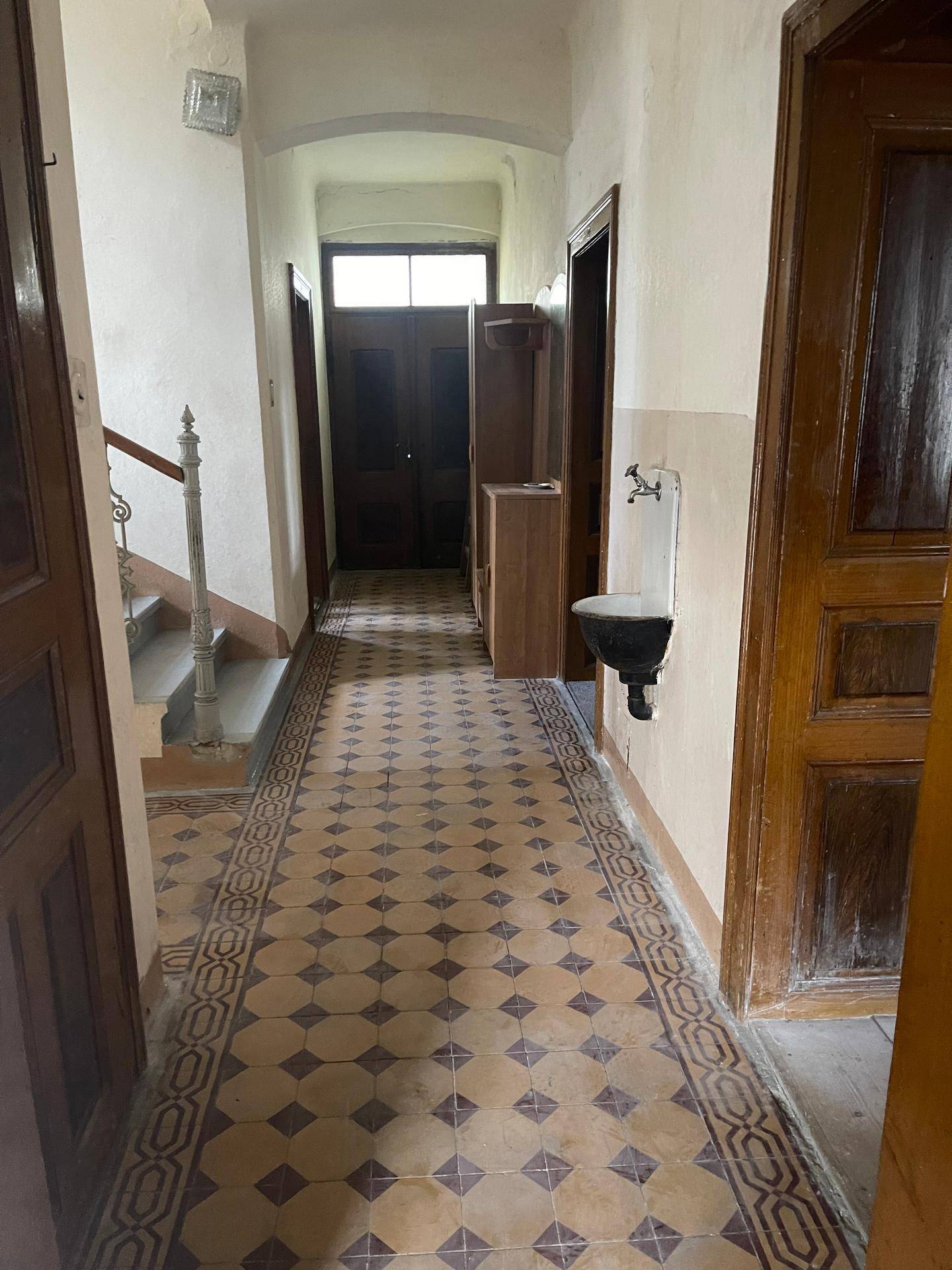 Langer Flur mit gemusterten Fliesen, Holztreppe und einem Wandwaschbecken in einem historischen Haus.