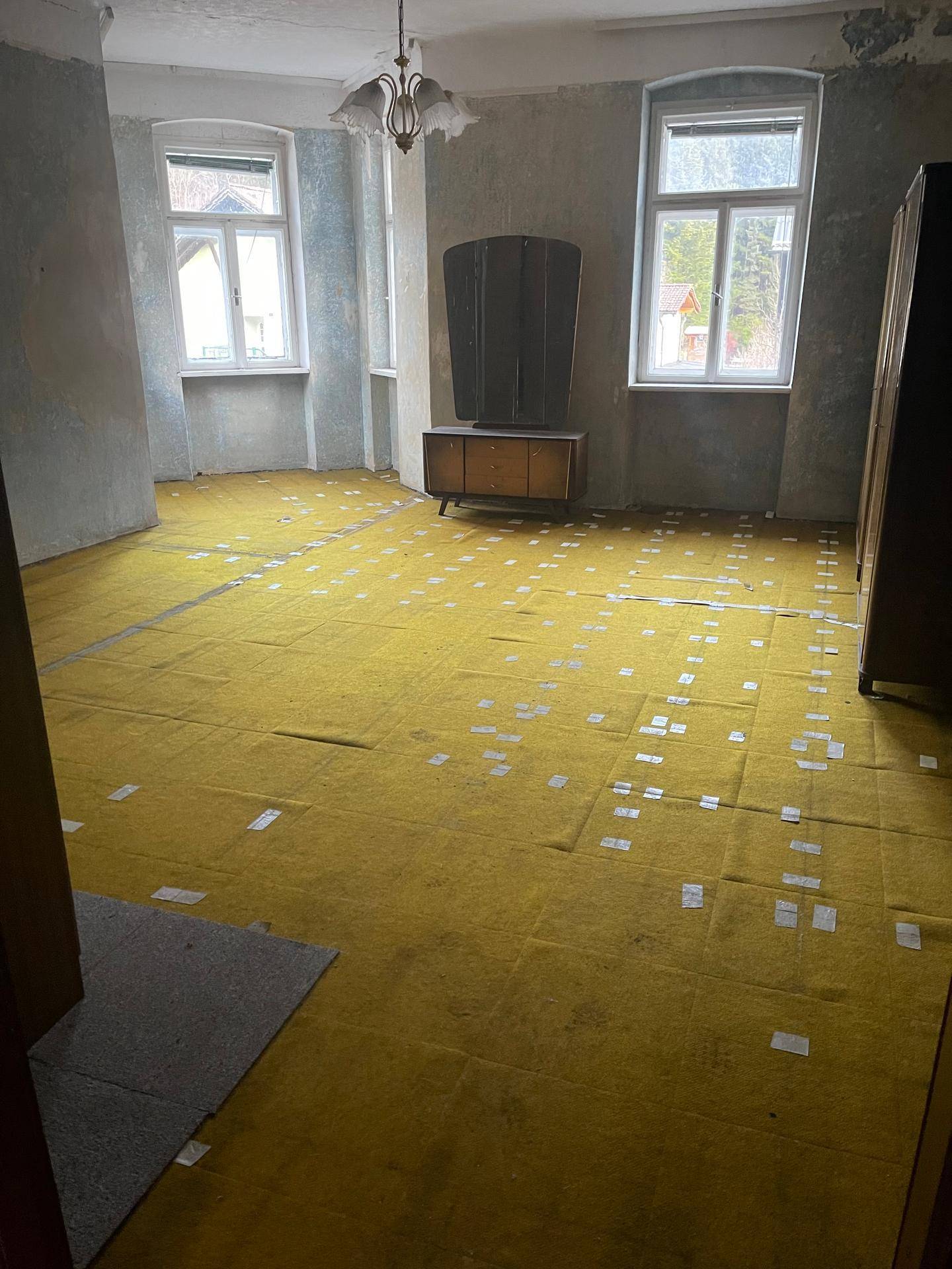 Geräumiges Zimmer mit gelbem Unterboden, zwei Fenstern und alten Möbeln, bereit zur Renovierung.