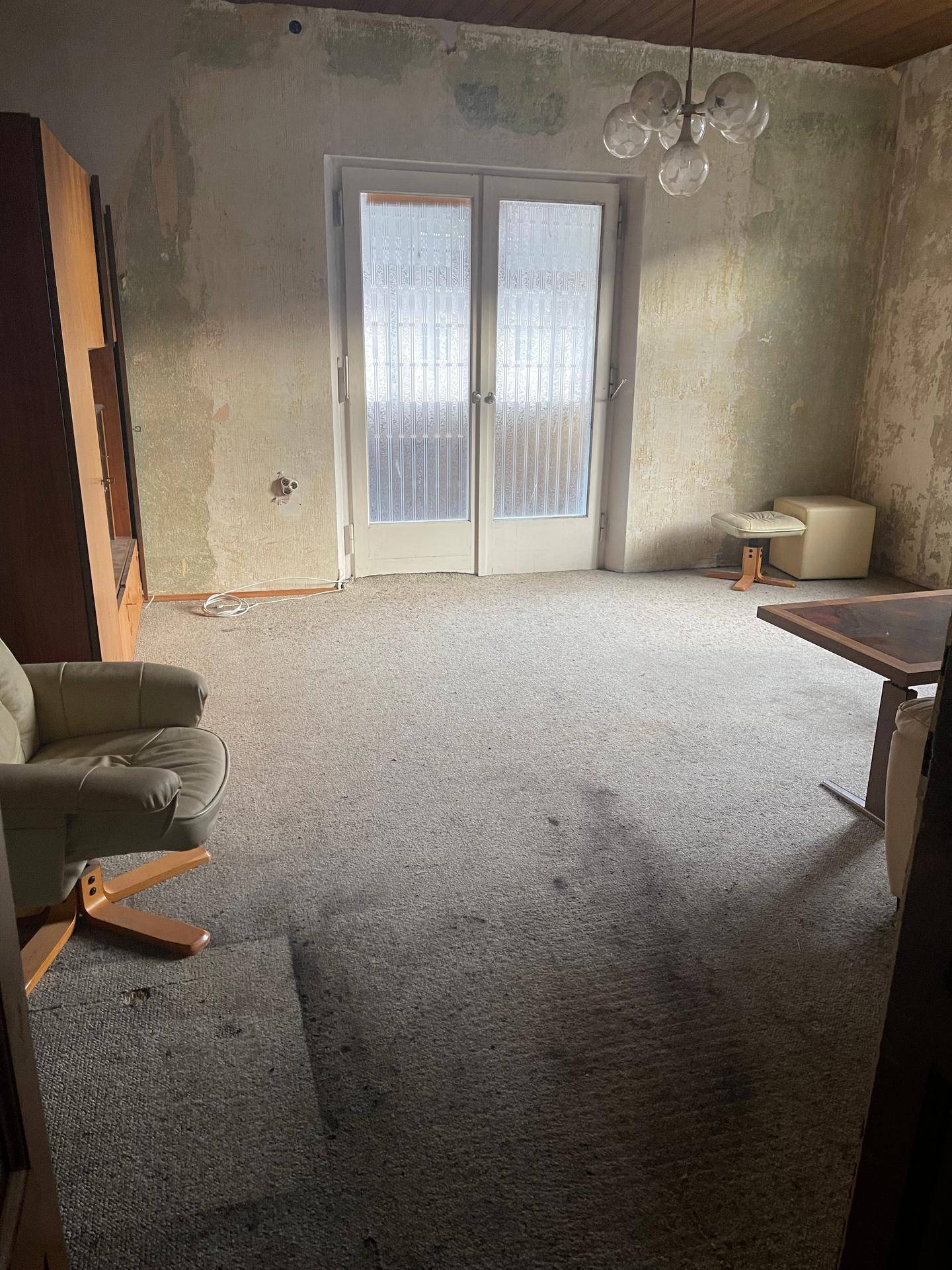Wohnzimmer mit Teppichboden, einer Terrassentür und alten Möbeln, in einem renovierungsbedürftigen Zustand.