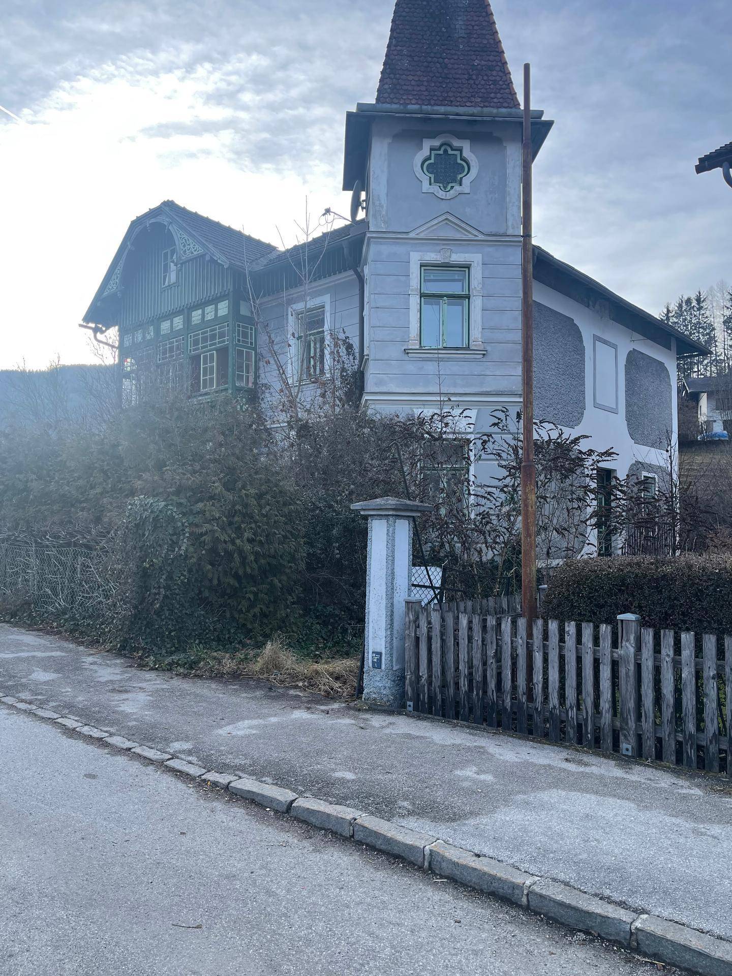 Historisches Gebäude mit Turm und Holzelementen, umgeben von Vegetation und einem Holzzaun.