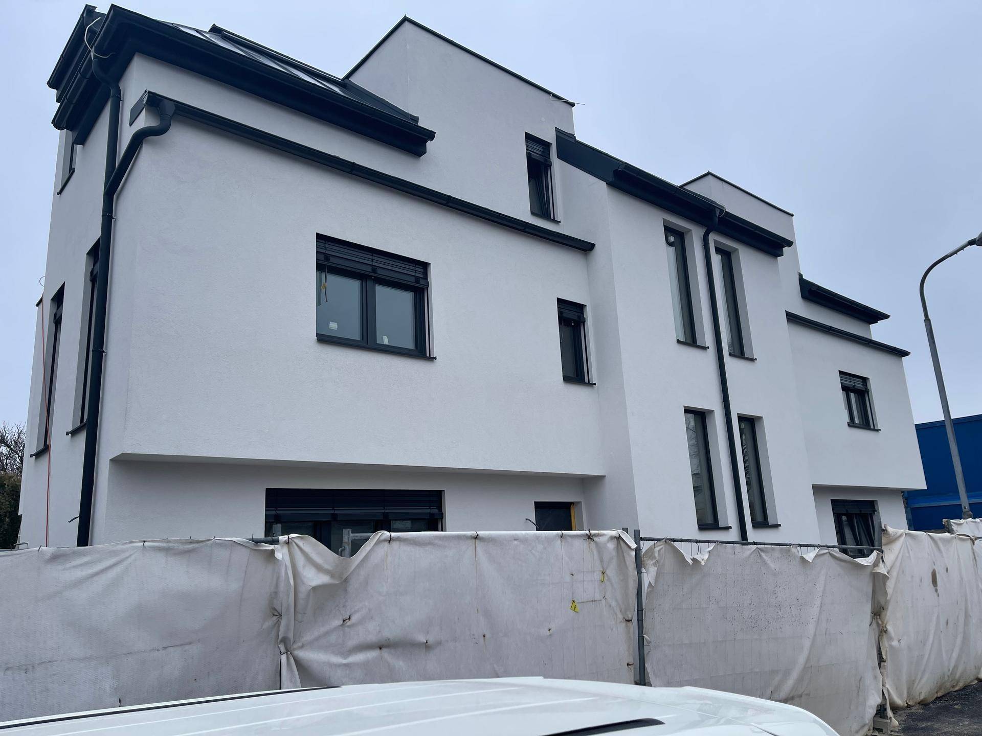 Ansicht eines modernen Neubaus mit weißer Fassade und dunklen Fensterrahmen, teilweise durch Bauzaun verdeckt.