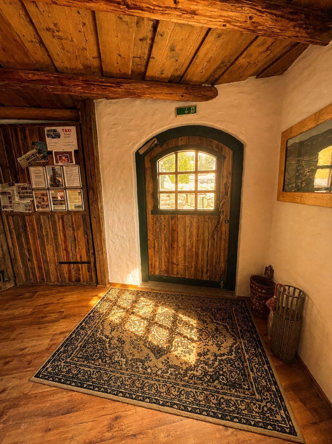 Detailansicht des rustikalen Eingangsbereichs mit Holztür, Teppich und Sonnenlicht, das auf den Holzboden fällt.