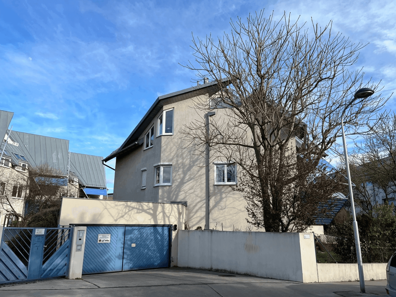 Fassade eines Wohnhauses mit blauer Garageneinfahrt, Zaun und kahlem Baum unter klarem Himmel.