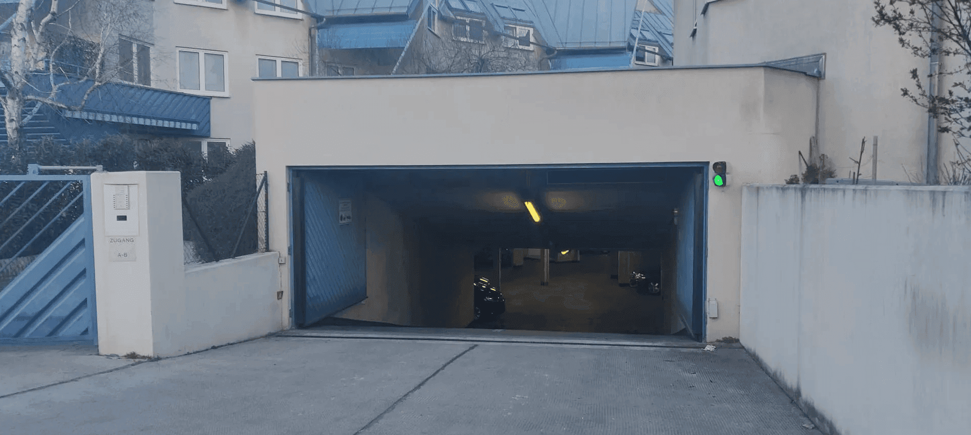 Offene Einfahrt zu einer Tiefgarage mit blauem Tor und grüner Ampel, umgeben von Wohngebäuden.