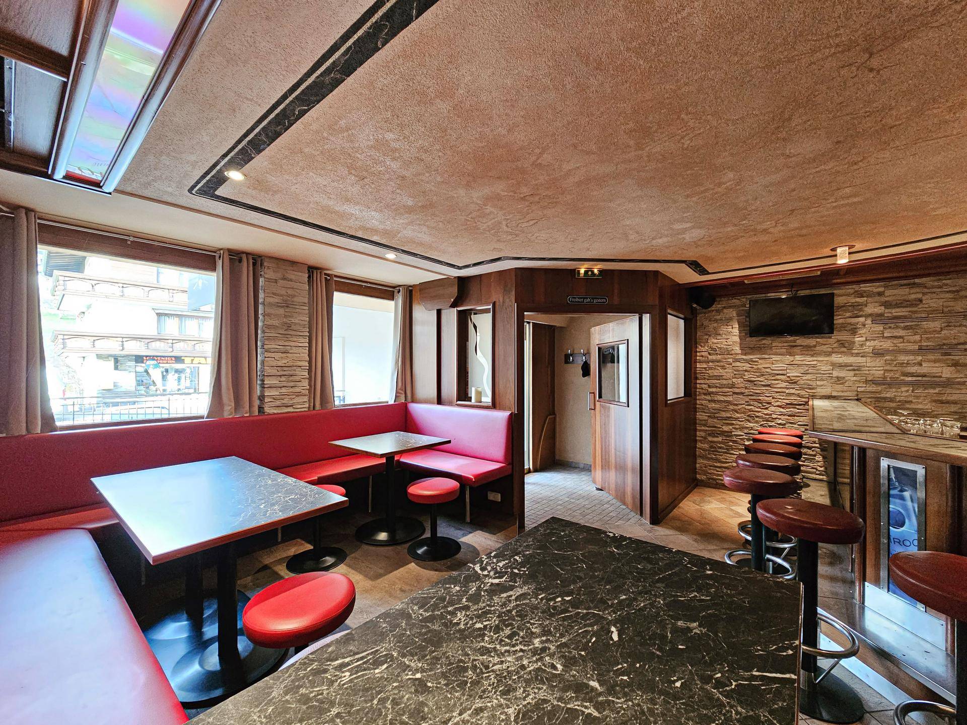 Gemütlicher Sitzbereich in der Bar mit roten Polsterbänken, Holztischen und Blick nach draußen durch Fenster.
