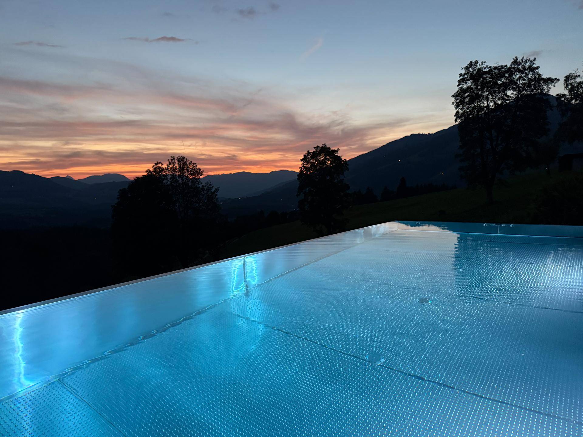 Beleuchteter Infinity-Pool bei Sonnenuntergang mit farbenprächtigem Himmel über den Bergen.