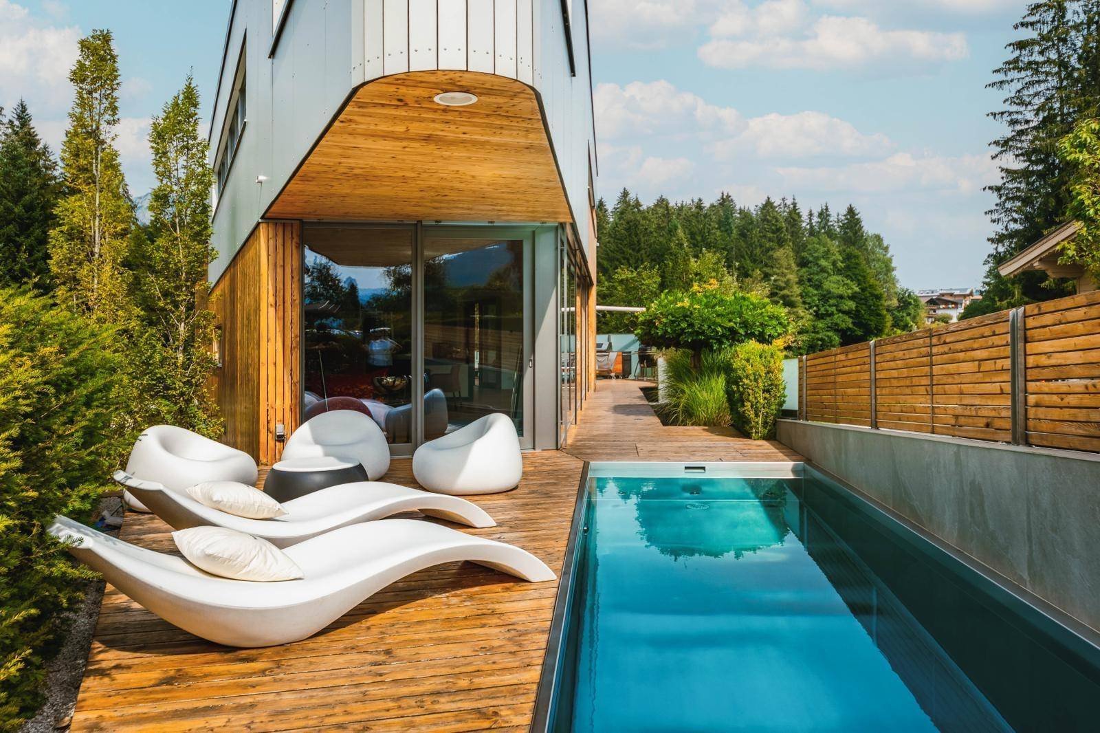 Moderner Außenpool mit Holzdeck und Liegen, umgeben von Hecken und Bäumen an einem sonnigen Tag.