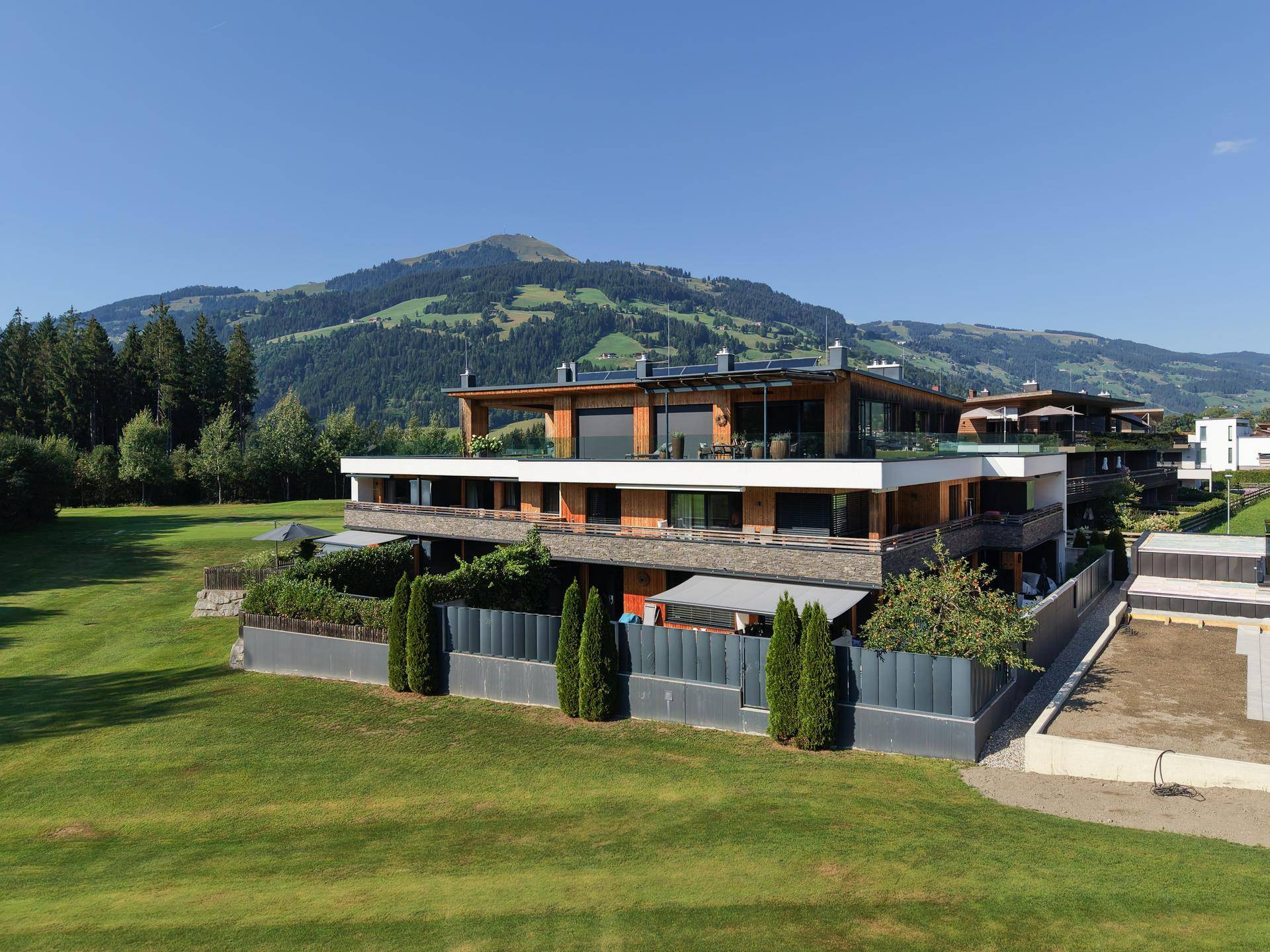 Modernes Wohngebäude mit Holzfassade und großen Balkonen, eingebettet in eine grüne Berglandschaft.