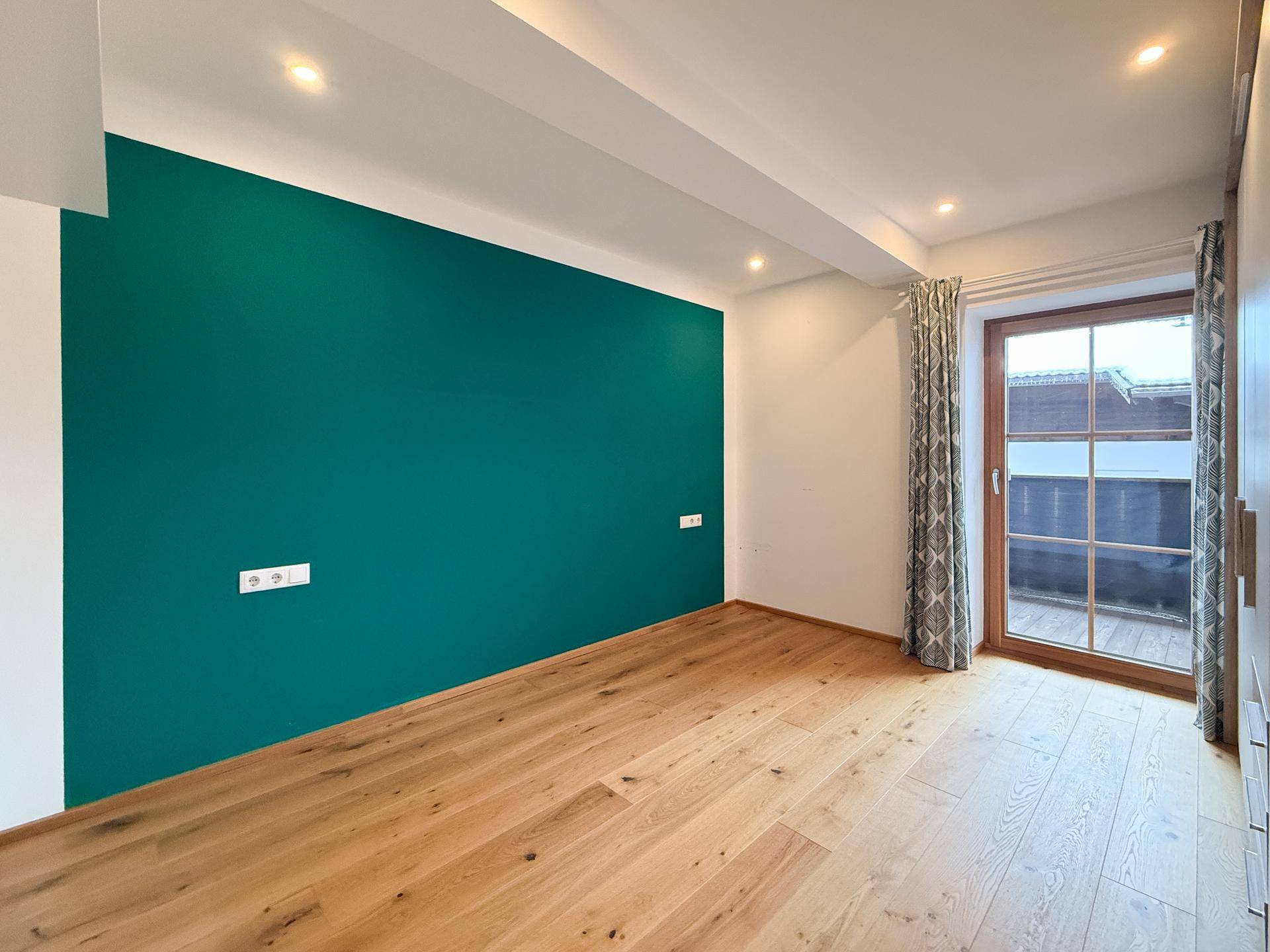 Schlafzimmer mit Holzboden, einer farbigen Akzentwand und Zugang zum Balkon.