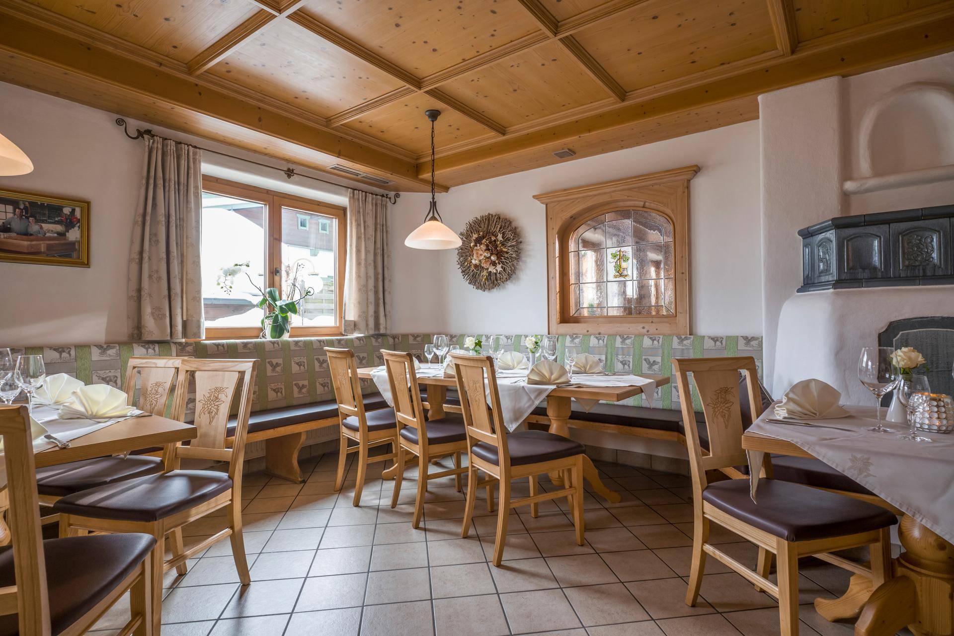 Heller Speisesaal mit Holzbänken, Tischen und einem traditionellen Kachelofen, der eine warme Atmosphäre schafft.