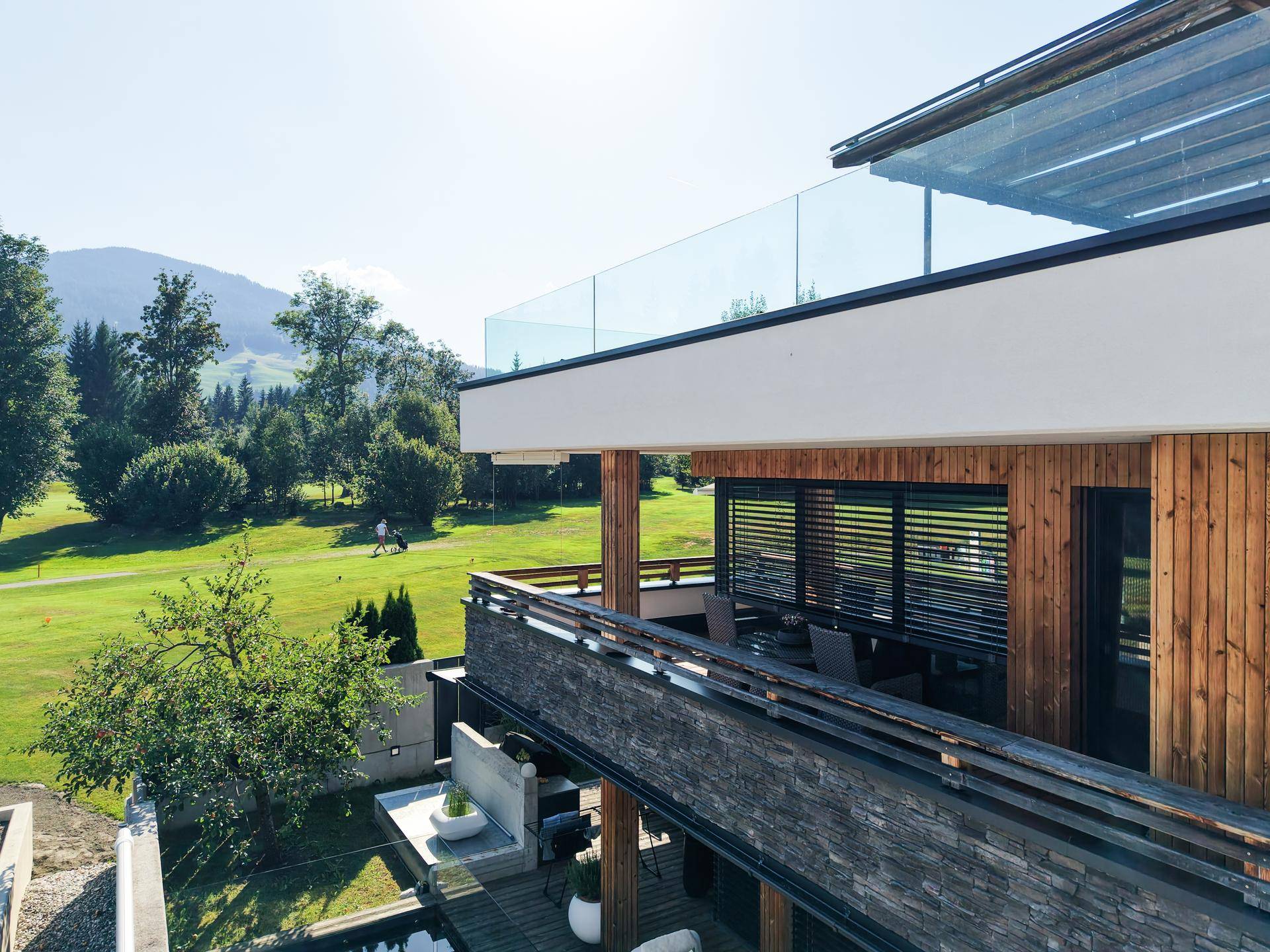 Geräumiger Balkon mit Holzboden und Steinverkleidung, umgeben von grüner Landschaft und Bergen.
