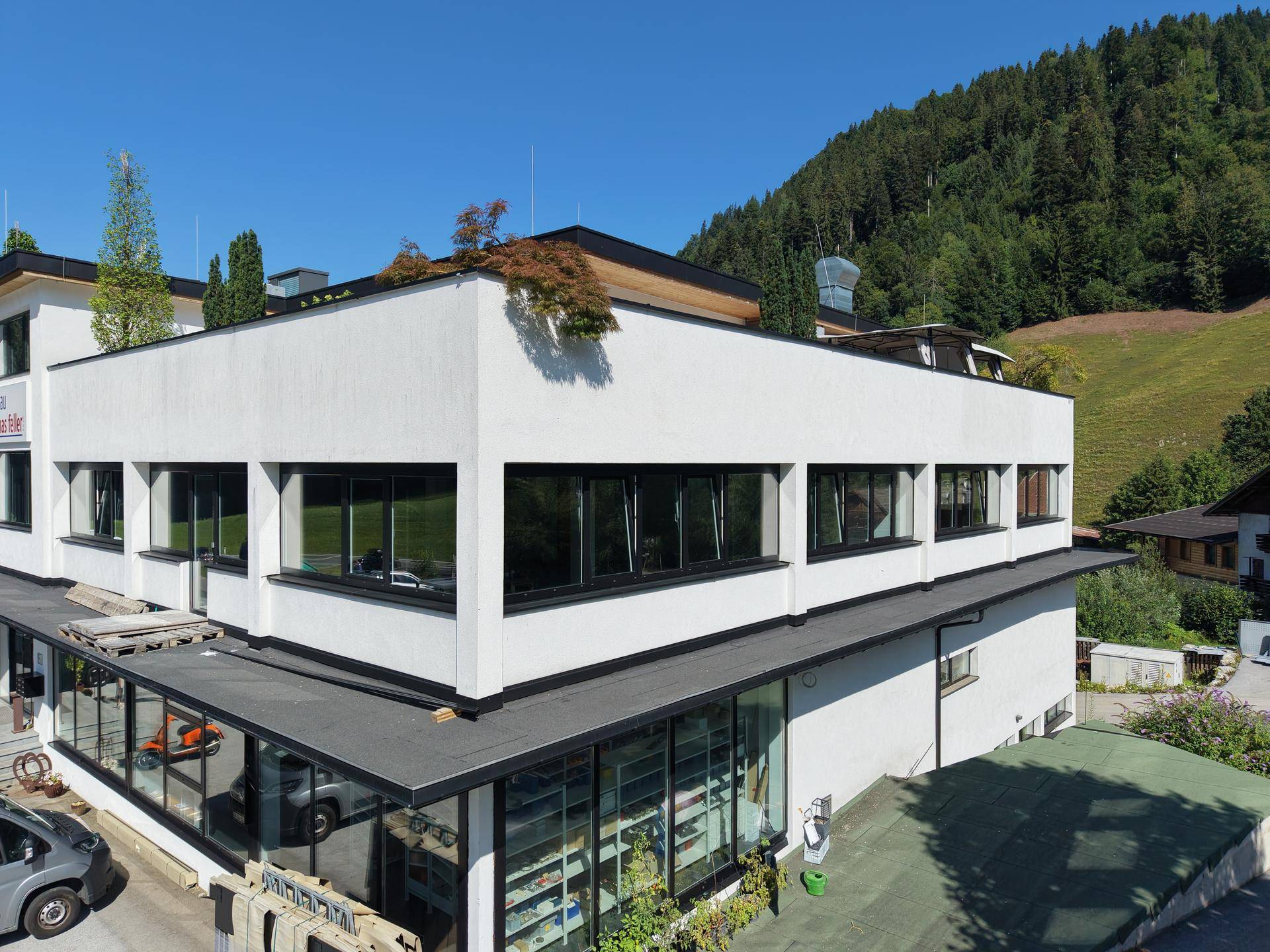 Moderne Gebäudefassade mit großen Fenstern und einem Flachdach, umgeben von grüner Natur und Bergen.