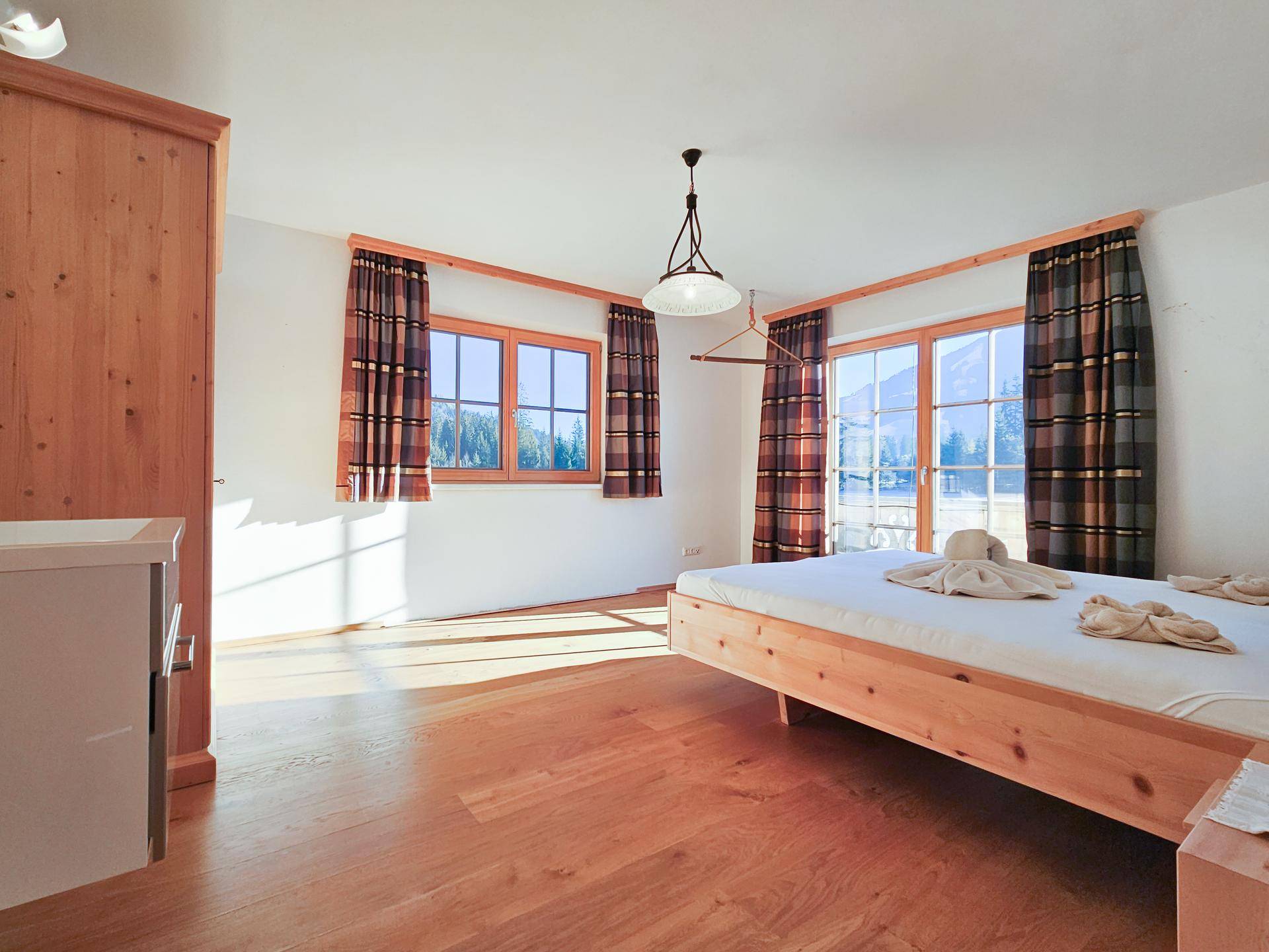 Helles Schlafzimmer mit Holzboden, einem einfachen Bettrahmen und großen Fenstern mit Zugang zum Balkon.