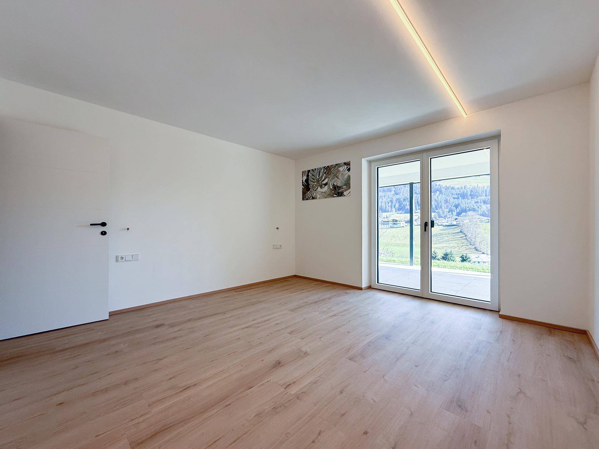 Heller, leerer Raum mit Holzboden und großem Fenster mit Blick ins Grüne.