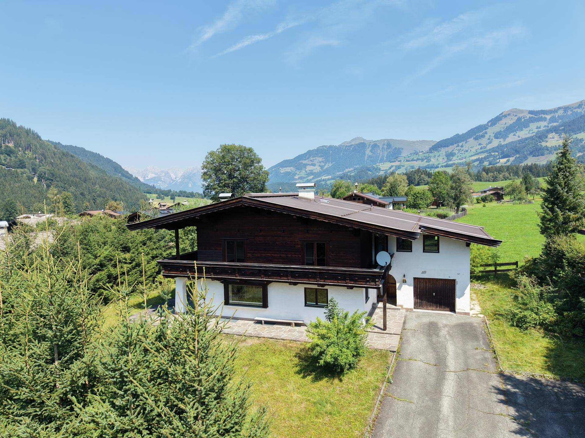 Haus im alpinen Stil mit dunklem Holzbalkon und Zufahrt, eingebettet in eine Berglandschaft.