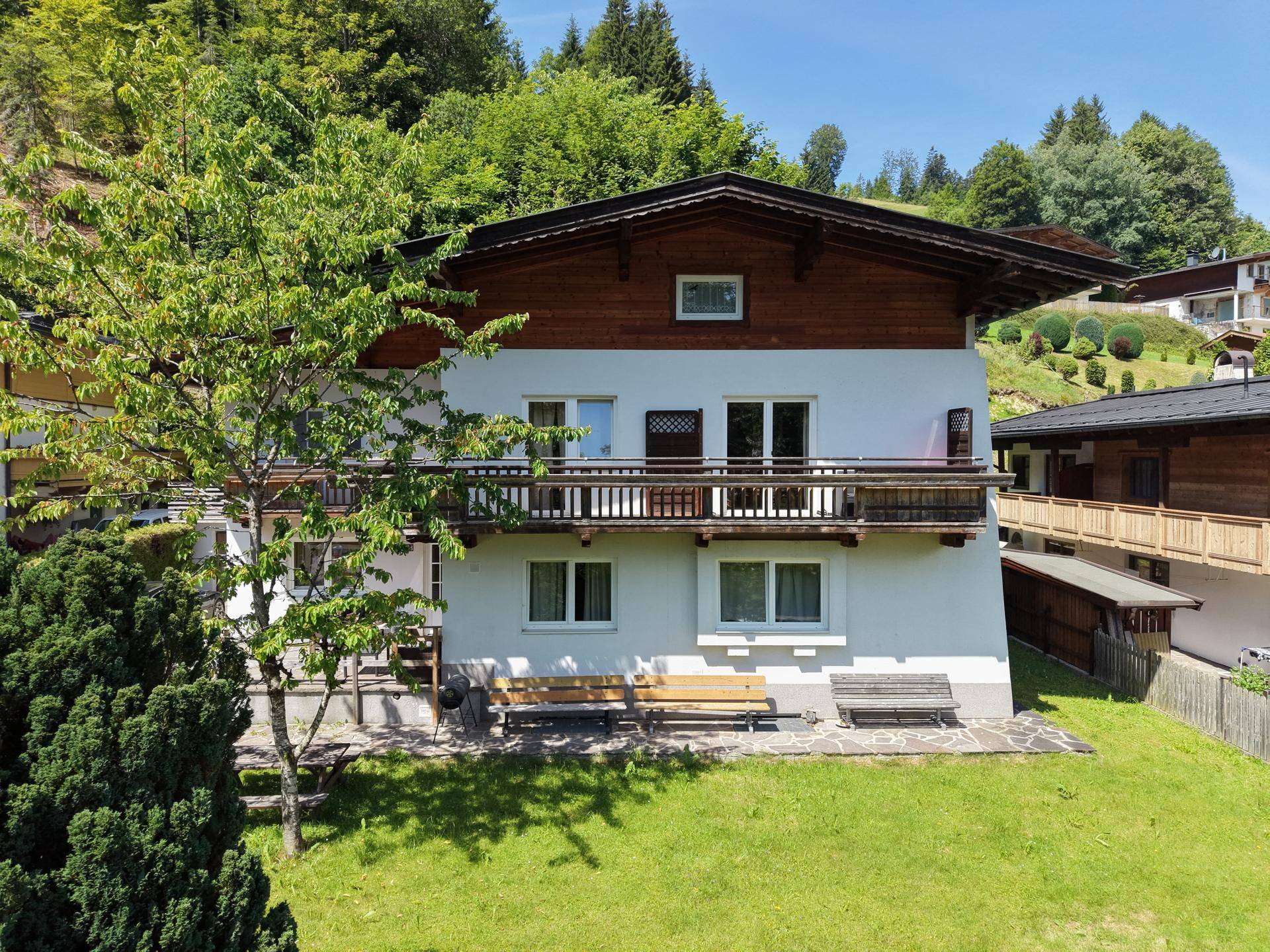 Vorderansicht des Hauses mit Balkon, gepflegtem Rasen und Sitzbänken im Grünen.