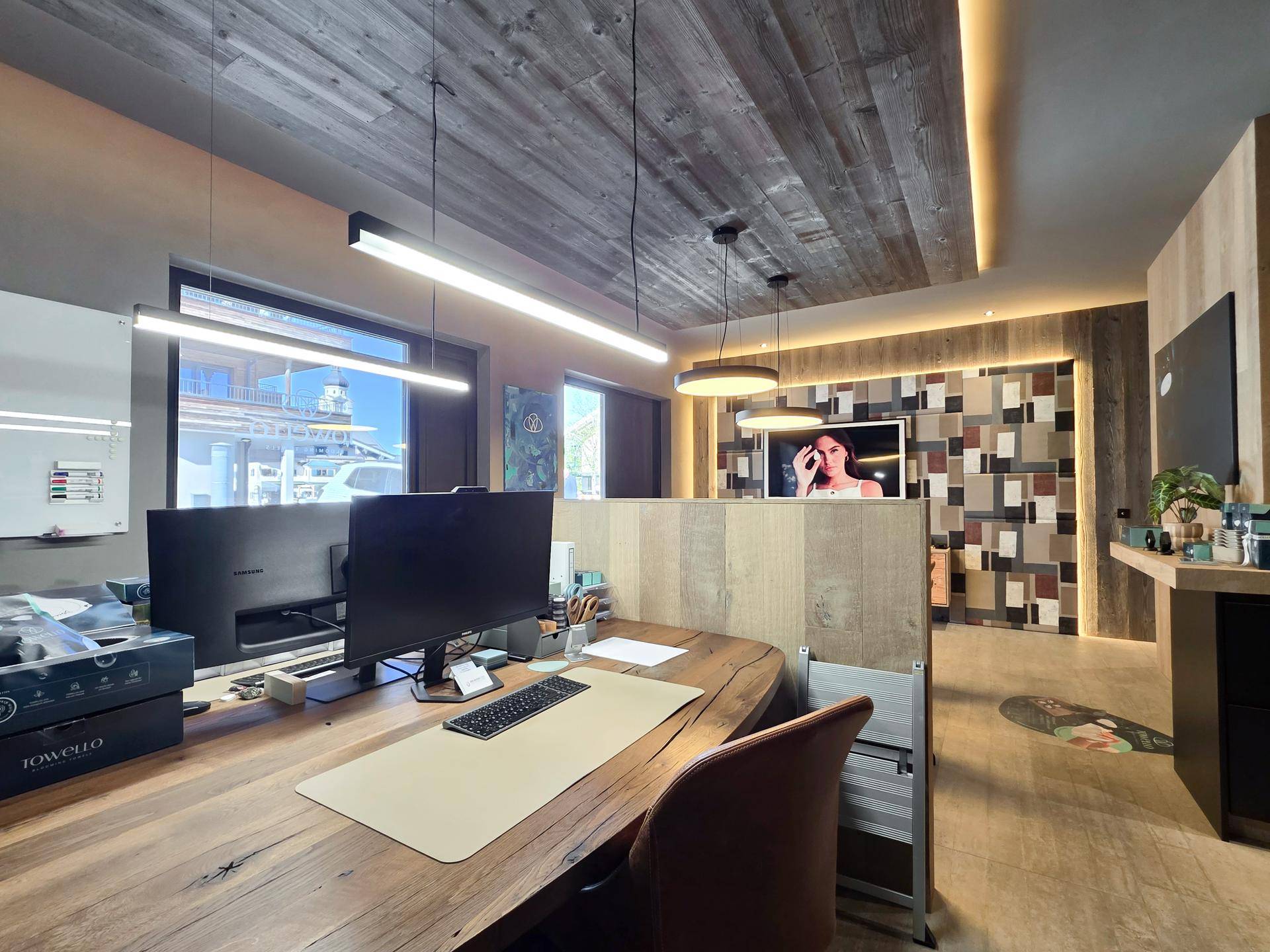 Arbeitsplatz in einem modernen Büro mit großem Holztisch, Computermonitoren und natürlichem Lichteinfall.