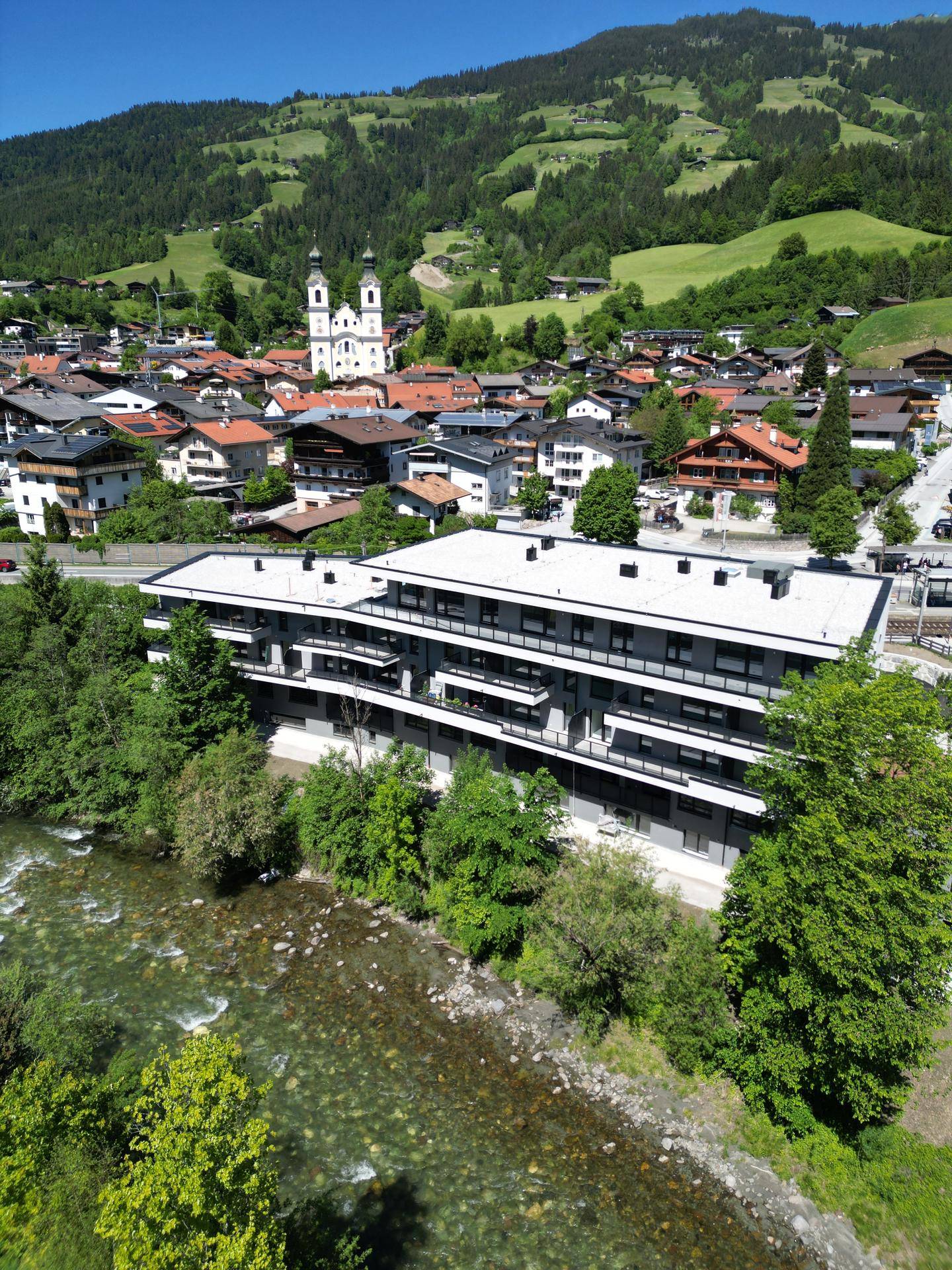 Luftaufnahme eines modernen Wohngebäudes am Flussufer mit Bergkulisse.