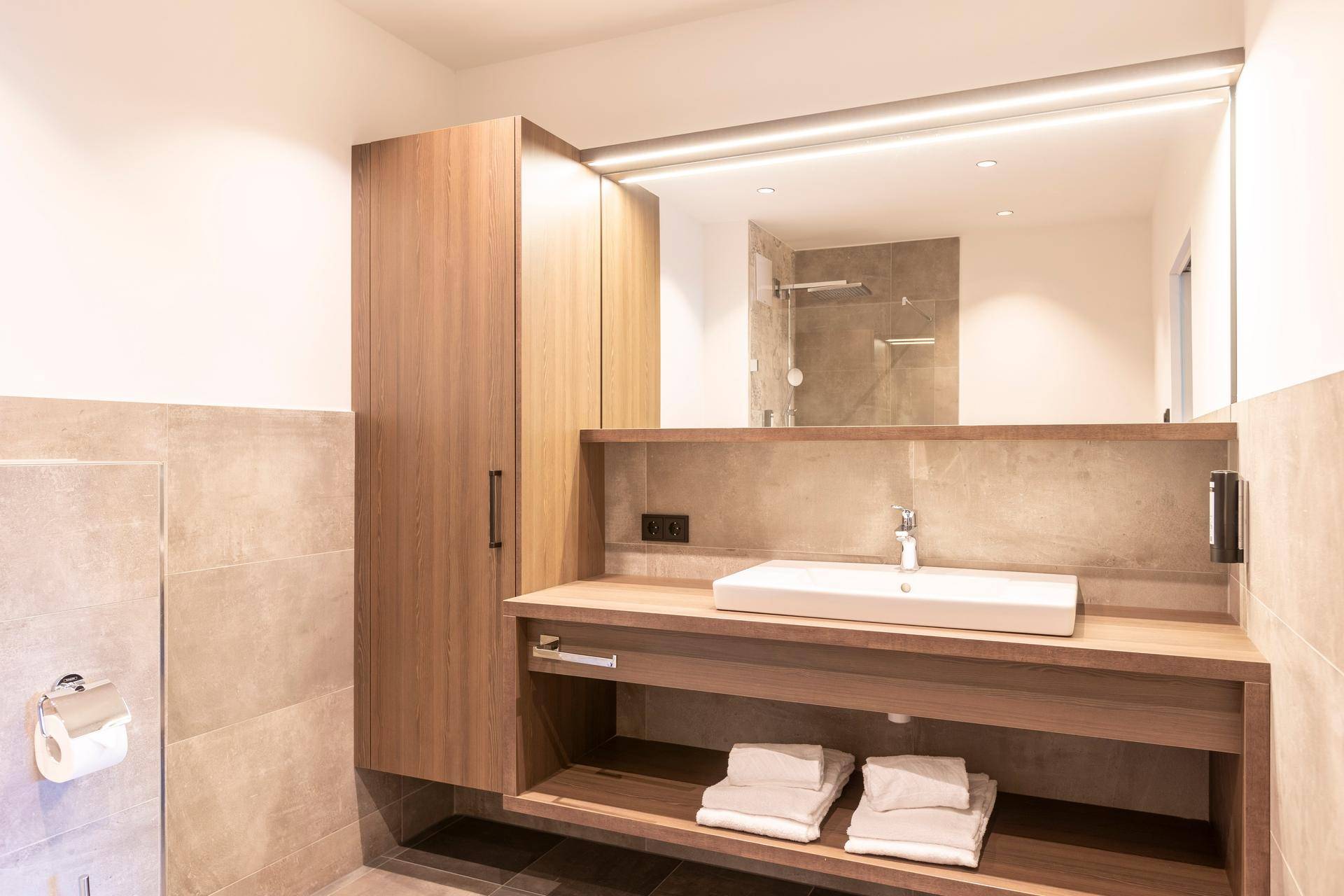 Helles Badezimmer mit modernem Waschbecken, beleuchtetem Spiegel und praktischem Holzschrank für Handtücher.