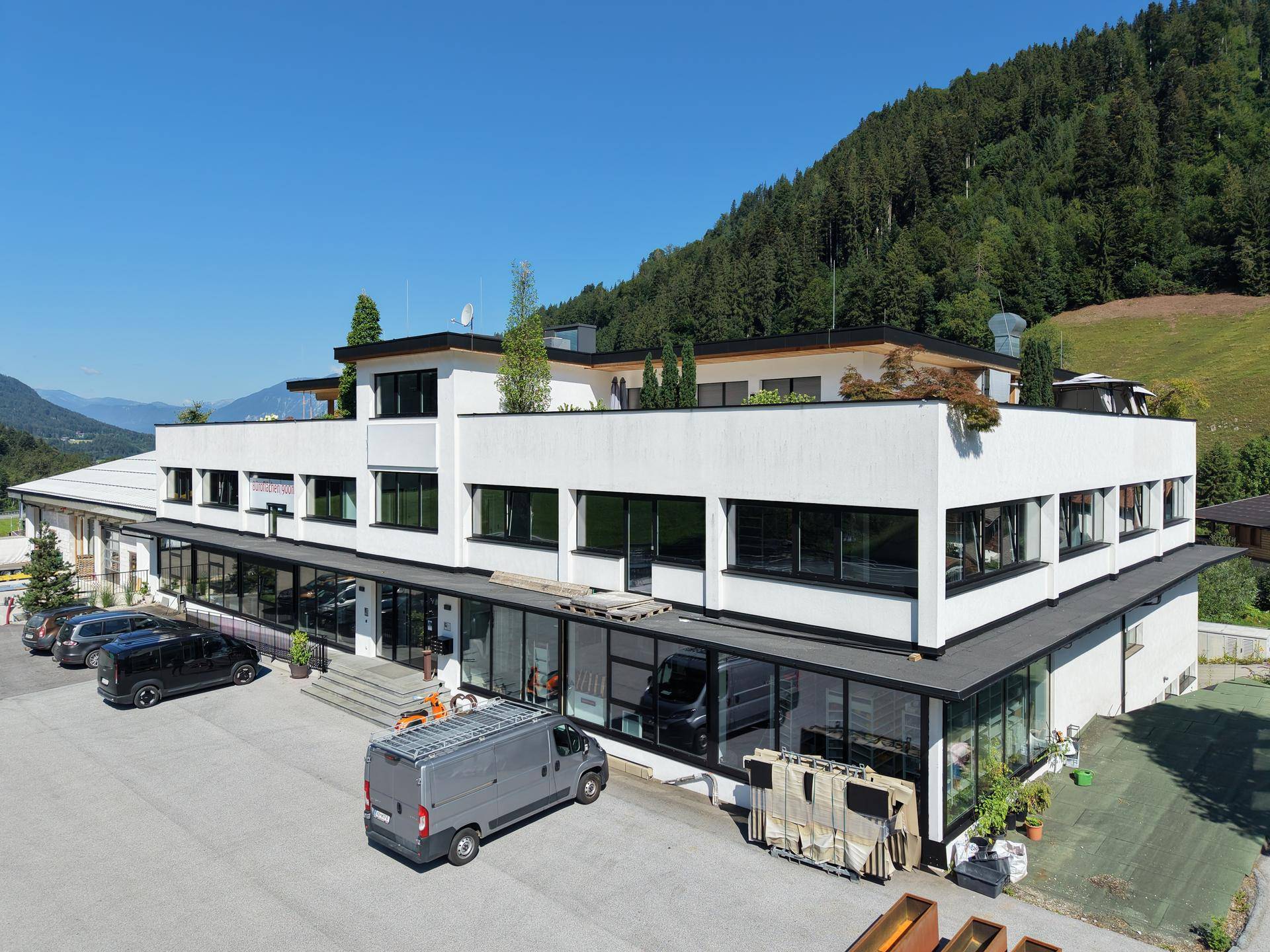 Modernes Geschäftsgebäude mit großen Glasfronten und Parkplätzen, umgeben von grünen Bergen.