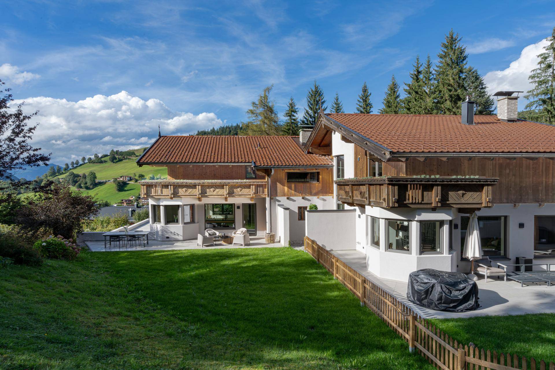 Großzügiges Alpenchalet mit weitläufigem Garten und mehreren Terrassen, umgeben von grünen Hügeln.